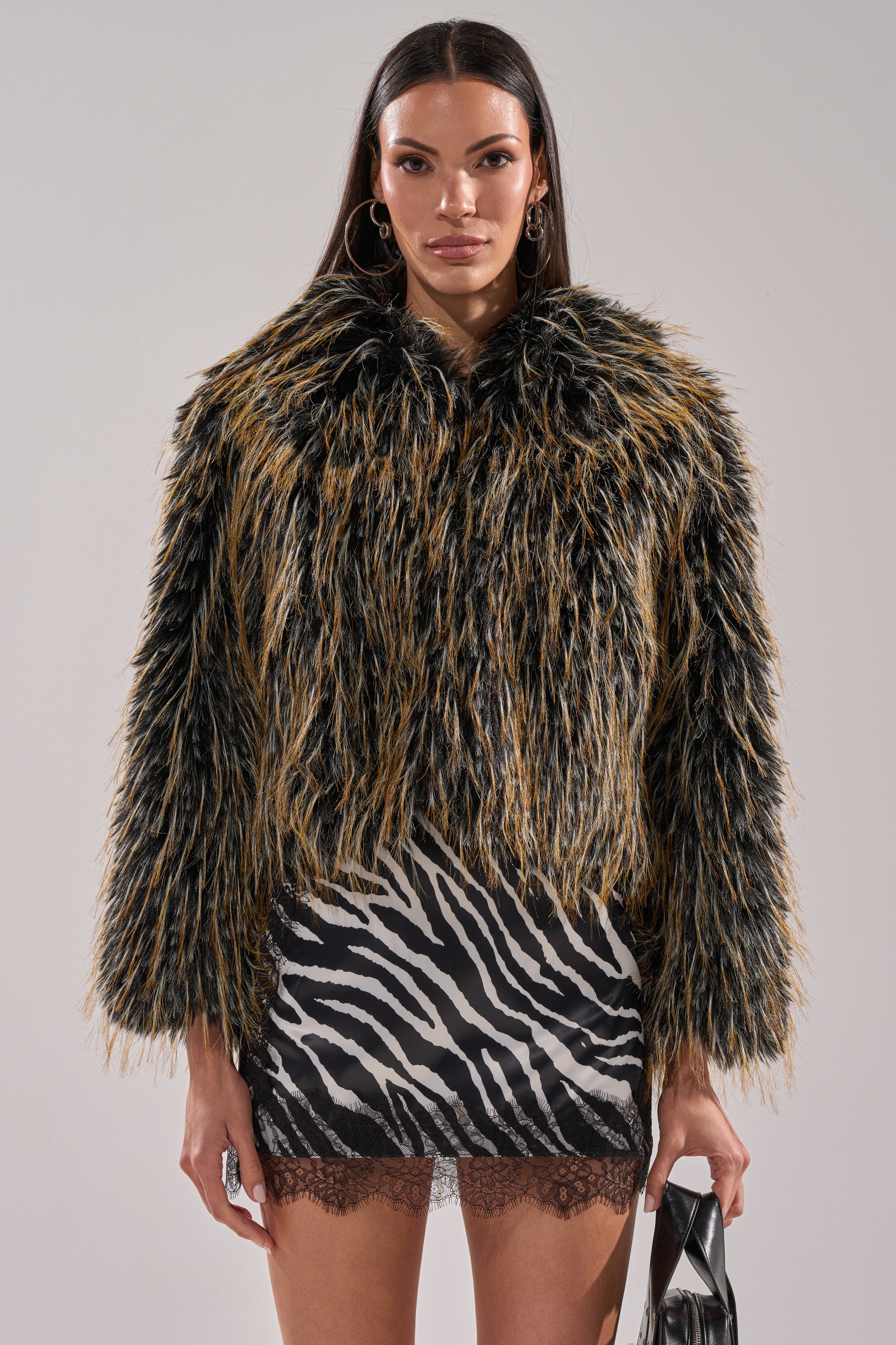 A woman faces forward, wearing the JUPITER FAUX FUR COAT—shaggy and multicolored with an oversized collar—over a zebra print mini skirt with lace trim. She holds a black purse and accessorizes with hoop earrings and long, straight hair.