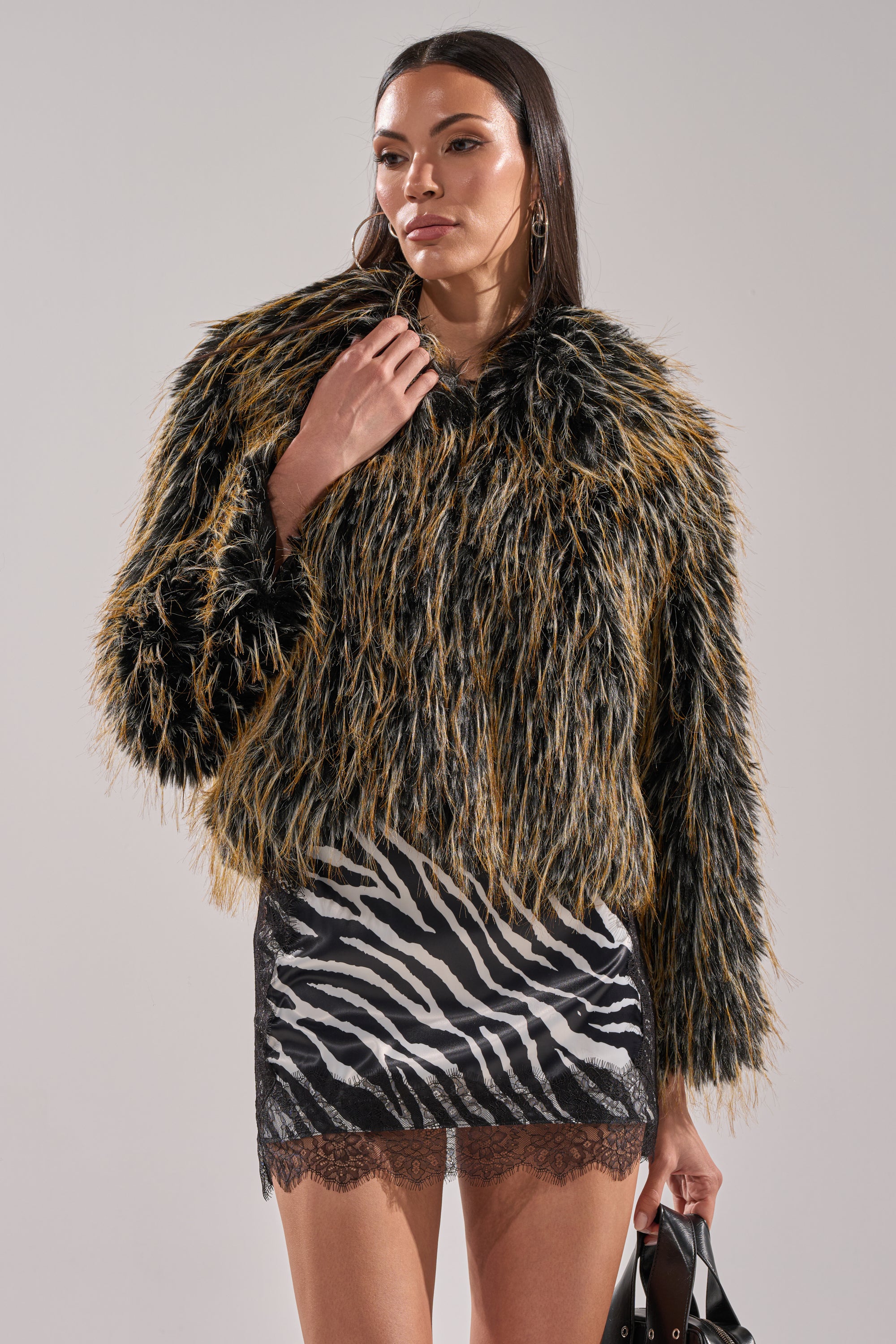 A woman confidently poses in the JUPITER FAUX FUR COAT—a shaggy brown and black piece with an oversized collar—over a zebra-print mini dress, accessorized with a black handbag and hoop earrings.