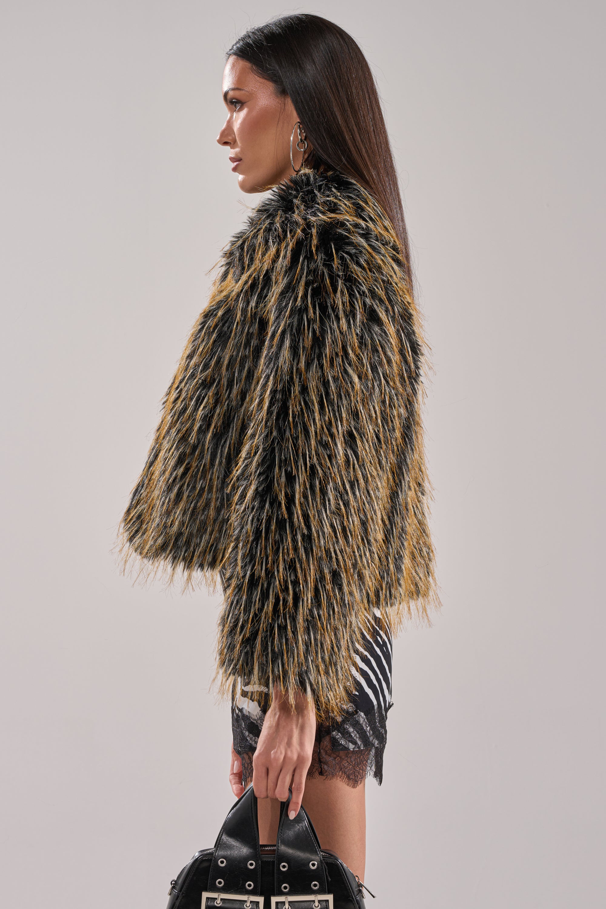 A woman with long dark hair stands in profile, wearing the JUPITER FAUX FUR COAT with an oversized collar, a black-and-white patterned skirt, large hoop earrings, and holding a black handbag with metal details.
