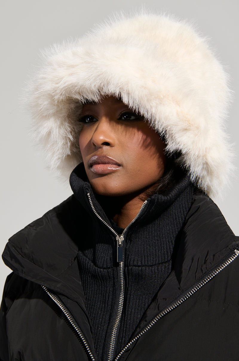 A person wearing a fluffy ivory faux fur bucket hat complements their black zip-up top, with the neutral background accentuating the cozy textures and chic winter style from the "BABY IT'S COLD OUTSIDE" collection by AKIRA Label.