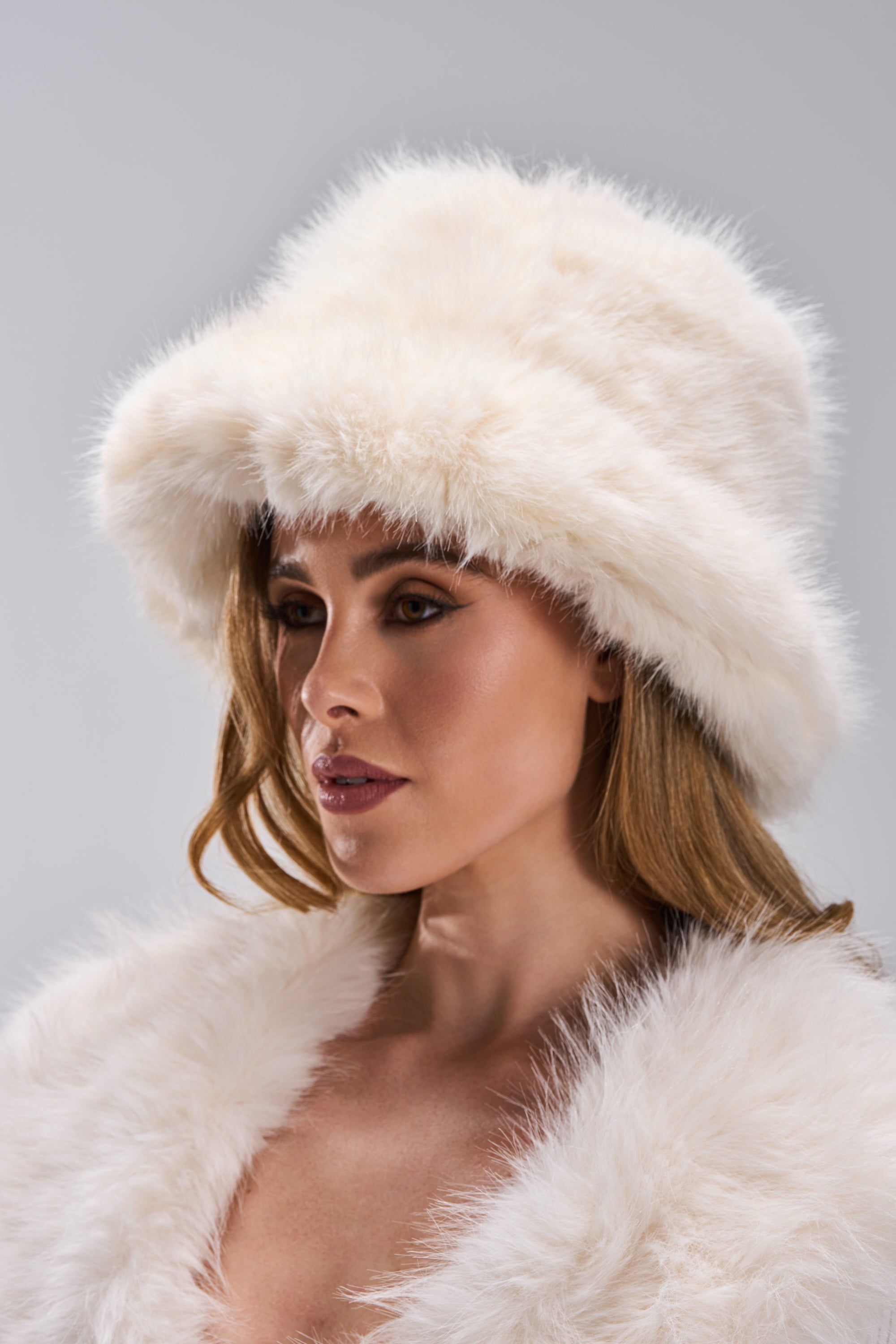 A woman with long light brown hair models the BABY IT'S COLD OUTSIDE FAUX FUR BUCKET HAT IN IVORY and a matching coat, looking to the side against a gray background—adding elegance to her winter wardrobe. 0
