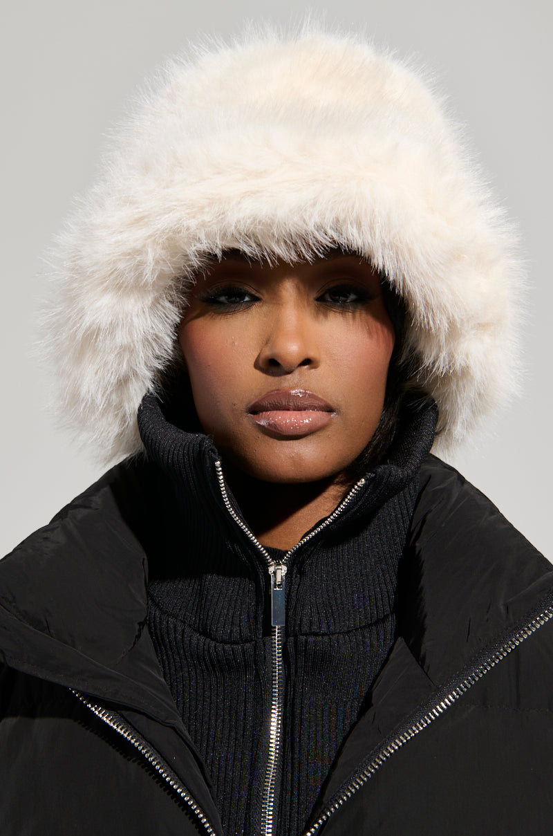 A person wearing the BABY IT'S COLD OUTSIDE FAUX FUR BUCKET HAT IN IVORY from the AKIRA Label gazes straight ahead. The plain, light-colored background accentuates their chic winter accessory.