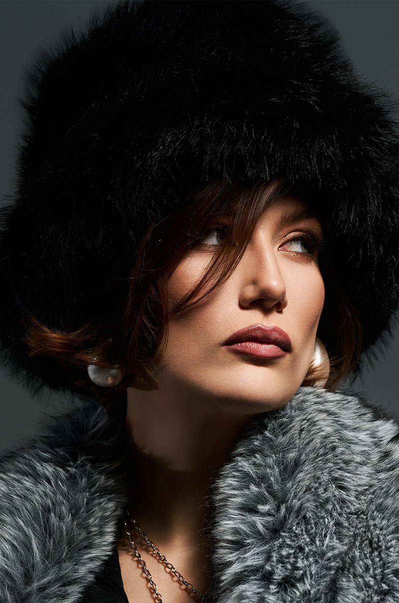 Dressed in stylish winter fashion, a woman wearing a gray coat and an AKIRA Label APRES SKI Faux Fur Bucket Hat in black gazes thoughtfully to the side. Her shoulder-length brown hair and pearl earrings enhance her look, while a subtle necklace adds a touch of elegance. The dark background accentuates her contemplative expression.