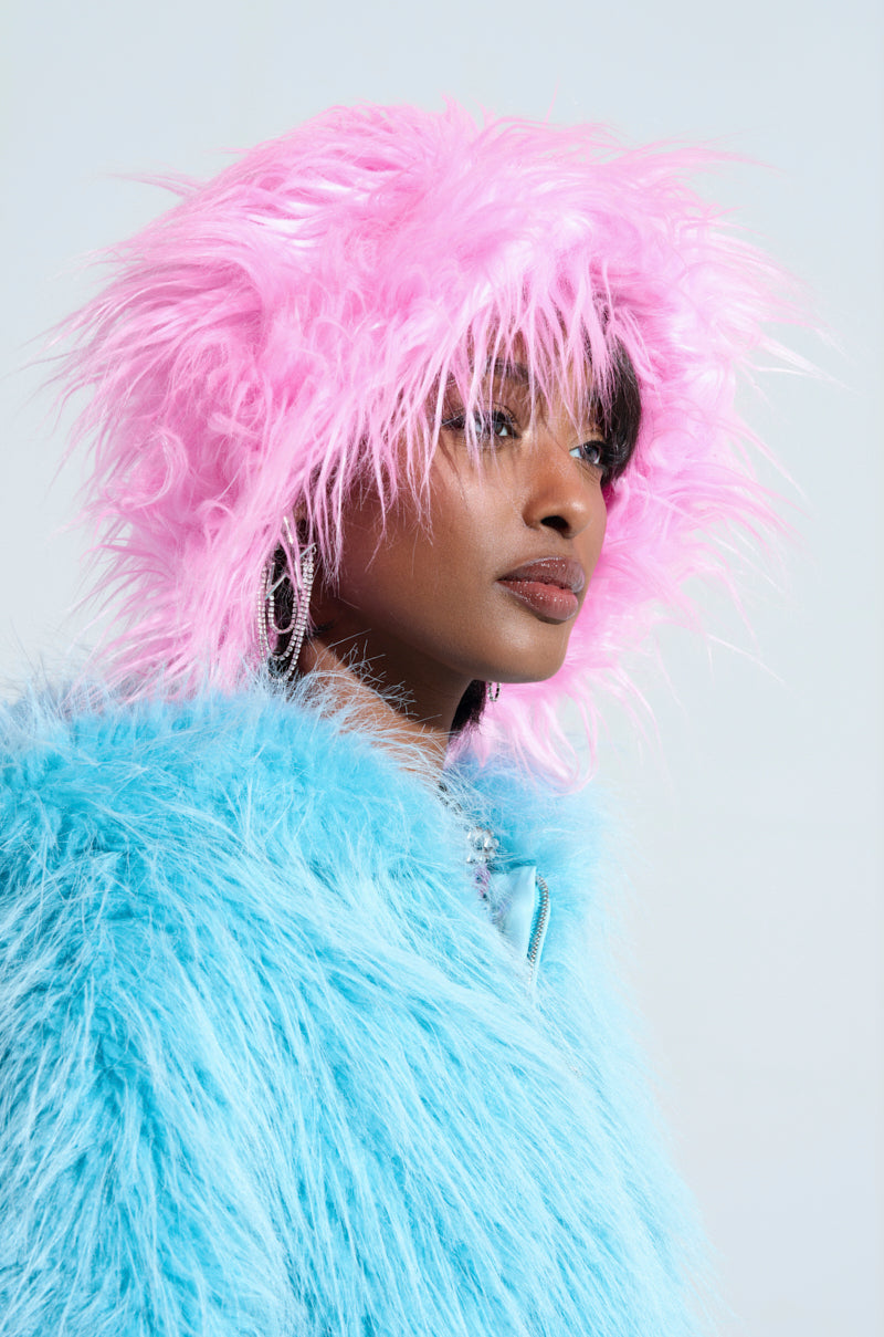 Wearing the I LOVE YOU FAUX FUR BUCKET HAT in vibrant pink with a fluffy texture, a person pairs it stylishly with a blue, shaggy coat. The hat perfectly complements their medium-length dark hair and sparkling earrings, offering an adjustable fit for comfort. Its eye-catching design stands out beautifully against the plain light background.