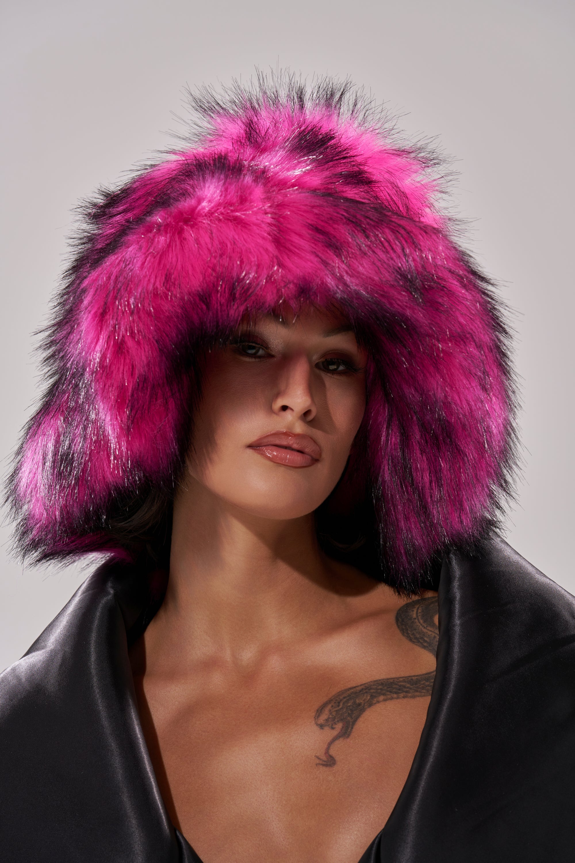 A woman wears the HOTTIE FAUX FUR BUCKET HAT, an oversized pink and black faux fur style, with a black coat. She showcases a snake chest tattoo while posing against a plain light gray background. 0