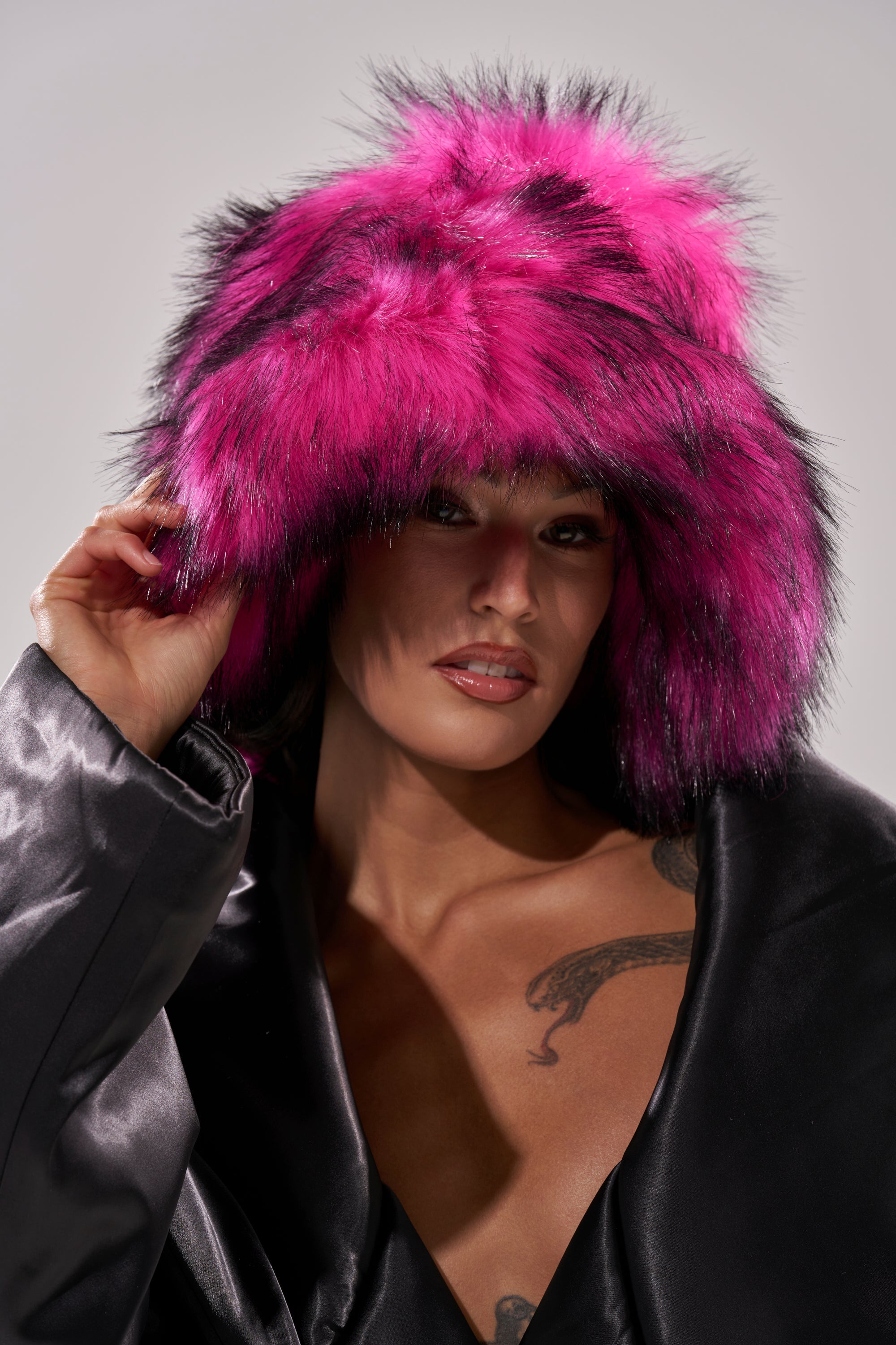 A woman poses confidently in the HOTTIE FAUX FUR BUCKET HAT, a vibrant pink accessory. She lifts the hat with one hand, showing off her shiny black jacket and revealing a striking snake tattoo on her chest. 1