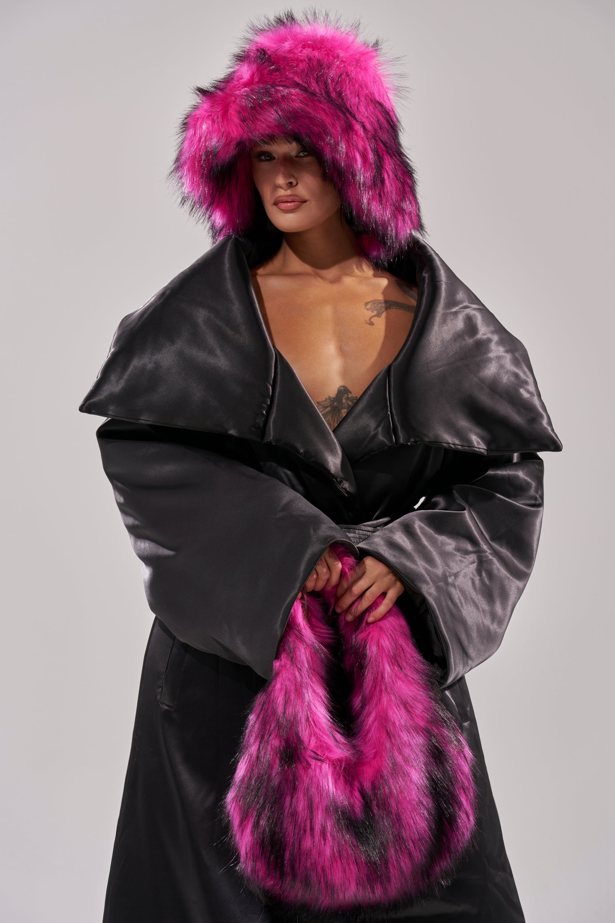 A person stands against a light gray background, wearing a black satin coat with oversized lapels, a matching hood and muff in vibrant pink and black faux fur, styled with the HOTTIE FAUX FUR BUCKET HAT. 2