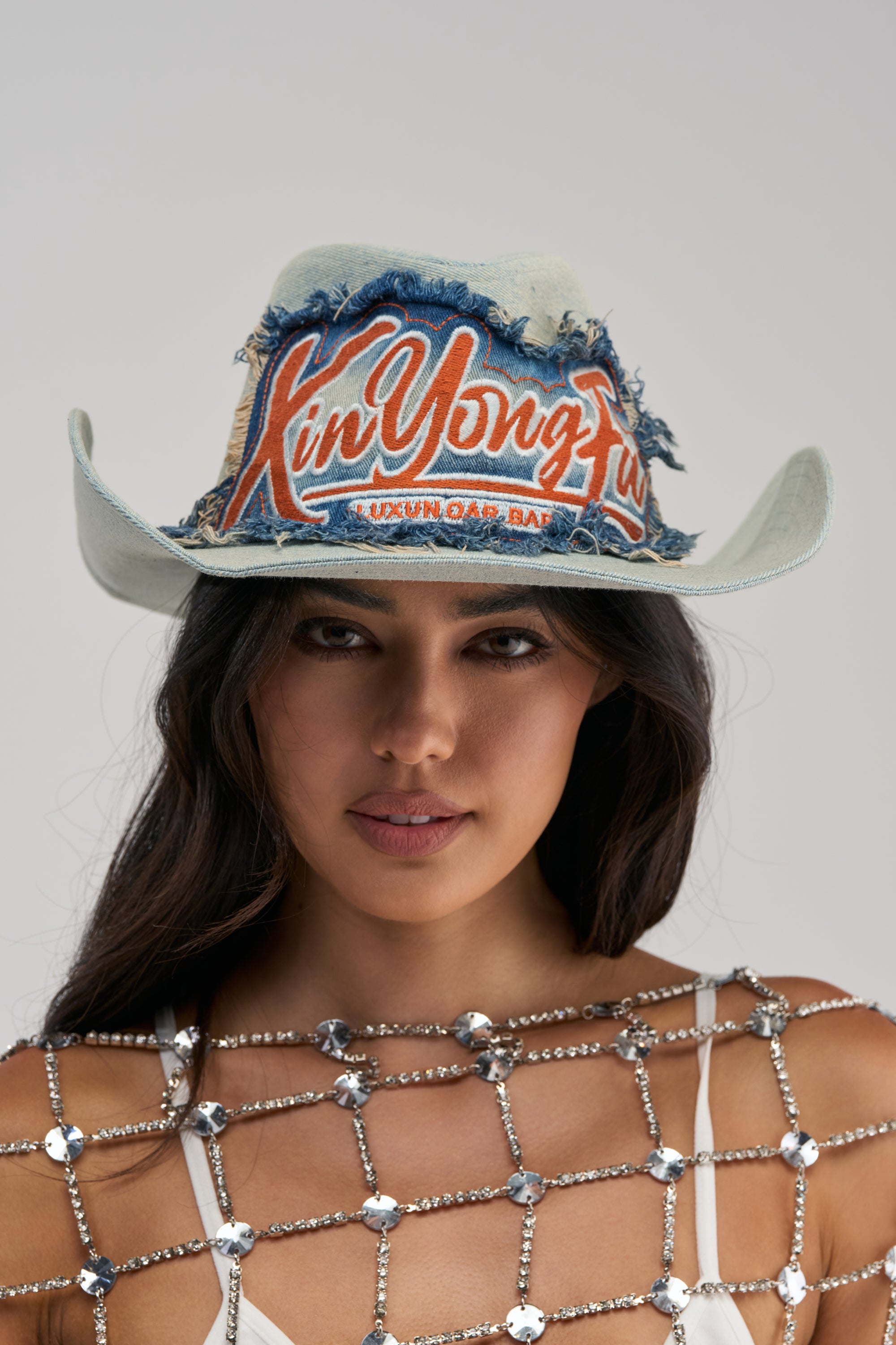 A woman wearing the WITH THE TIMES DENIM COWBOY HAT—featuring distressed denim, frayed edges, and “Xin Yong Lu” in bold orange lettering—paired with a rhinestone-studded mesh top layered over white, poses against a simple backdrop for an effortlessly western-inspired festival look.