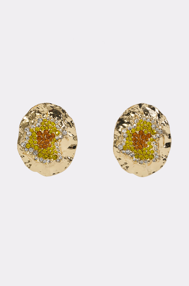 The GOLDEN HOUR EARRING IN RUBY is a pair of gold, round, textured statement earrings adorned with clusters of small yellow, orange, and clear gemstones at the center, displayed on a plain white background.