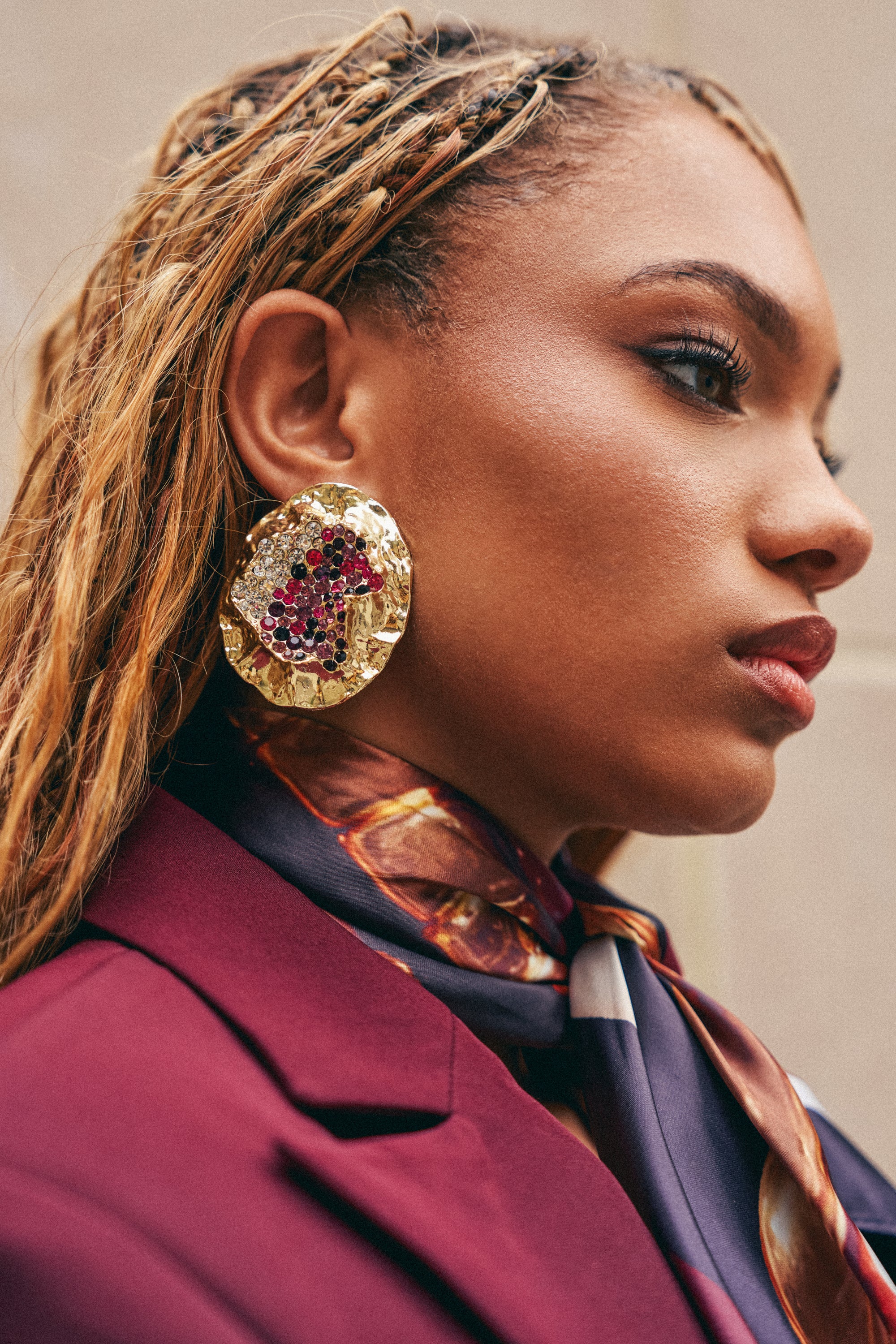 Wearing the GOLDEN HOUR EARRING—a bold statement piece featuring a rhinestone center and vivid red gemstones—a woman with braided hair pairs it with a burgundy blazer and a patterned scarf, gazing thoughtfully into the distance.