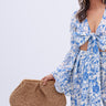 A woman with long gray hair stands against a white background, wearing a blue and white floral outfit with flared sleeves and holding the OUT OF OFFICE OVERSIZED RAFFIA CLUTCH, which features a woven raffia exterior.