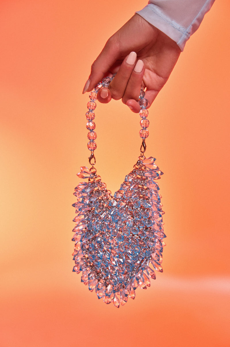 A manicured hand holds the ON MY TIME EMBELLISHED PURSE, featuring a crystal-inspired, spiky texture that channels mermaid glam against a vibrant orange background.