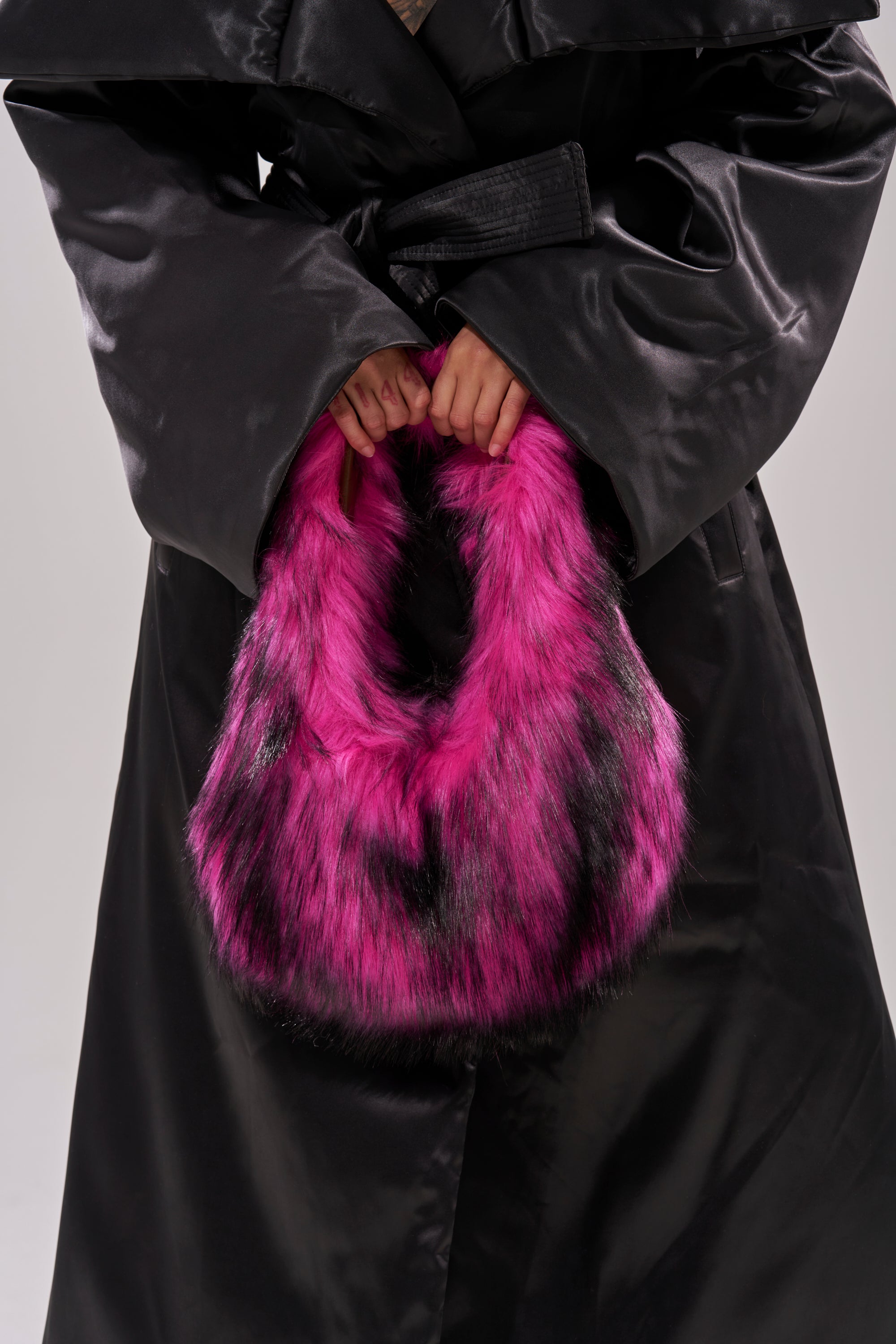 A person in a shiny black trench coat holds the IN NYC FAUX FUR PURSE—a fluffy, bright pink and black faux fur handbag—against a plain background.