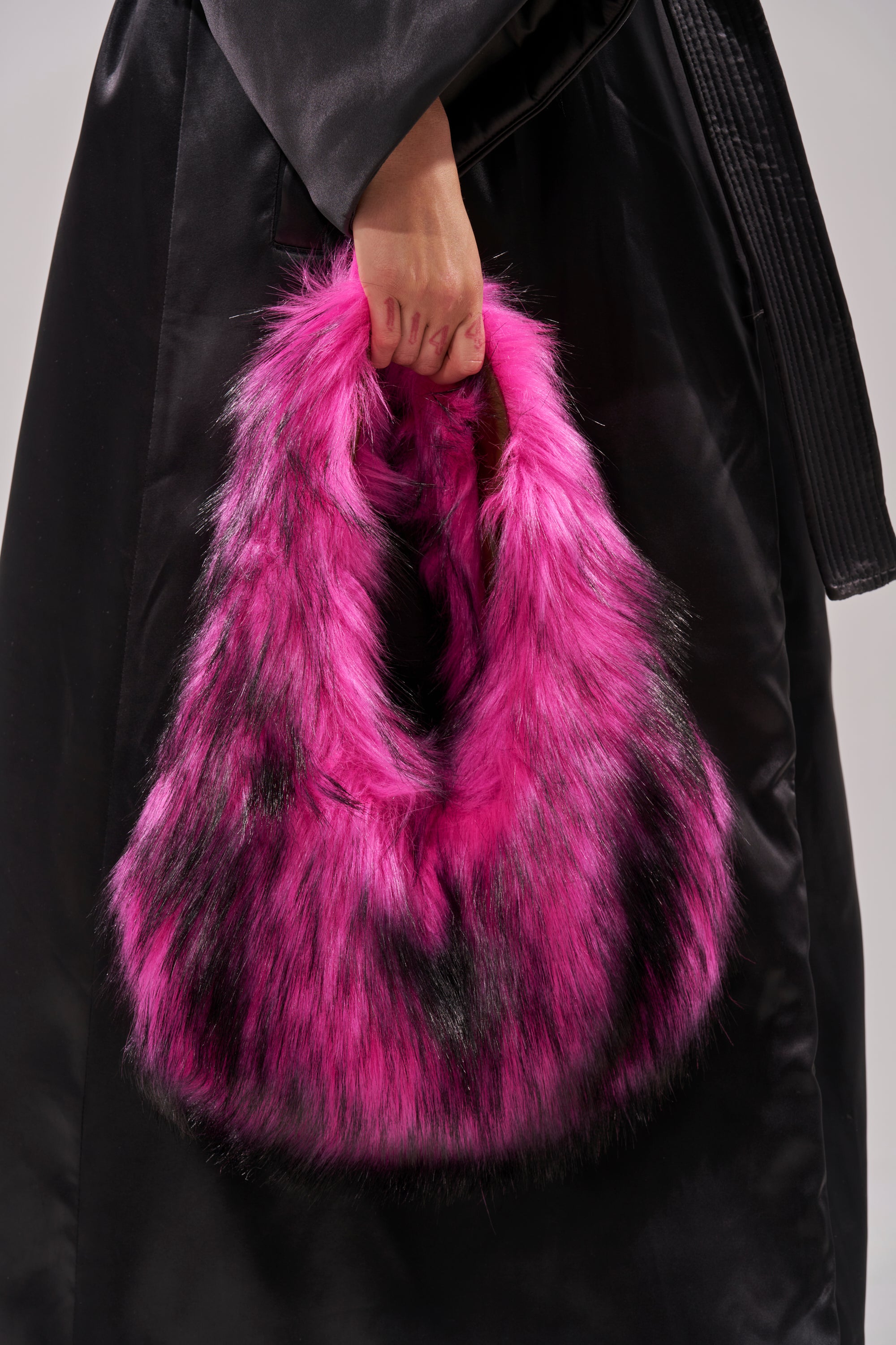 A person in a black coat holds the IN NYC FAUX FUR PURSE, a large, fluffy handbag made of vibrant pink faux fur with streaks of black.