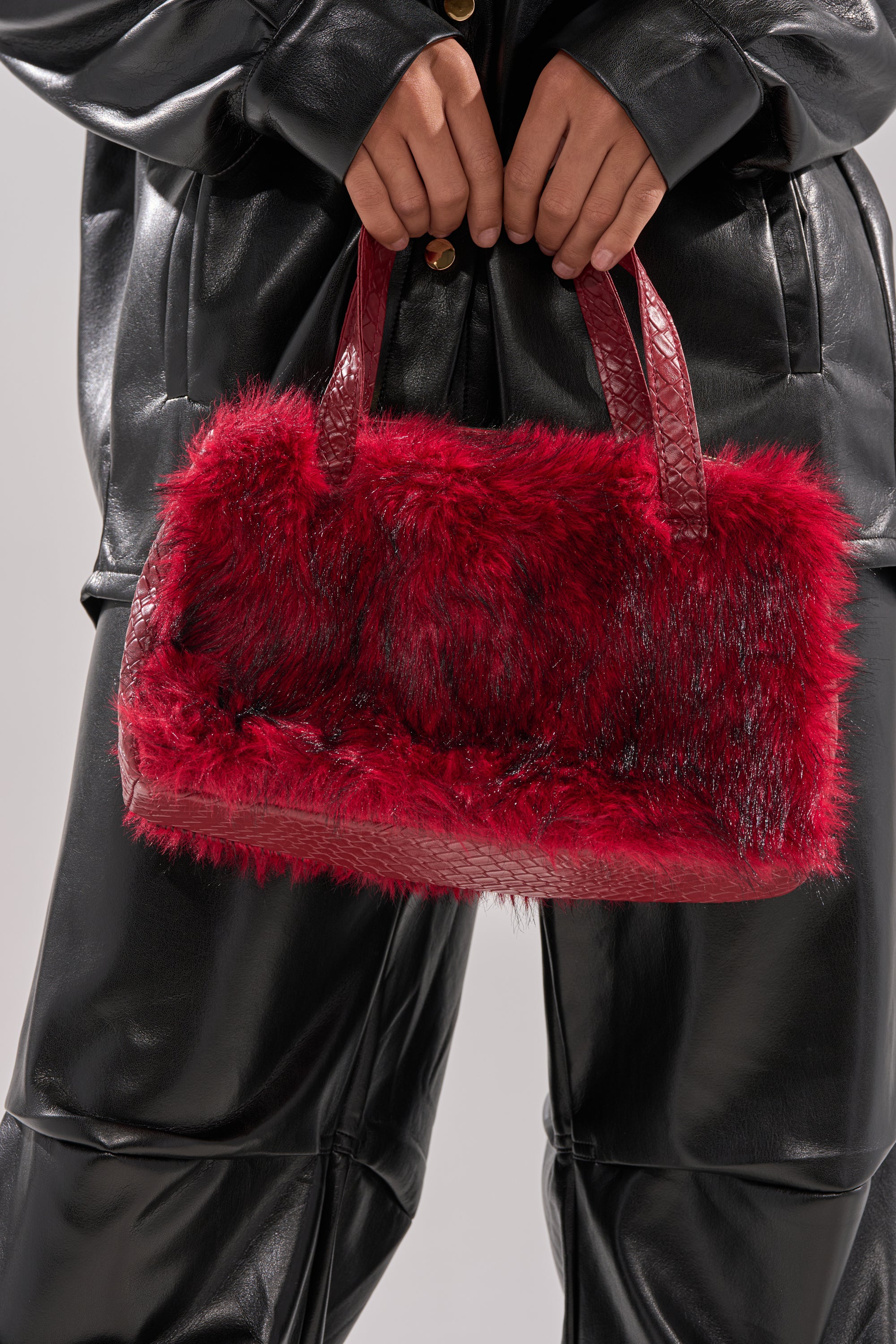 A person in a black leather outfit holds the TRUDY FAUX FUR PURSE, a red bag with two handles and snakeskin-embossed trim, in front of their body.