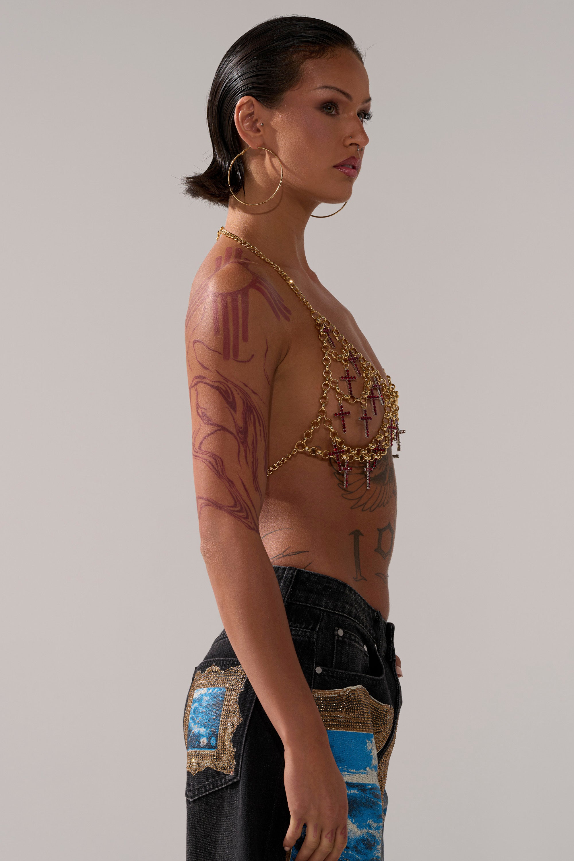 A person with short dark hair, hoop earrings, and visible tattoos poses in profile wearing the HOLY CROSS BODY CHAIN over a halter top, paired with black jeans featuring blue and gold patchwork details.