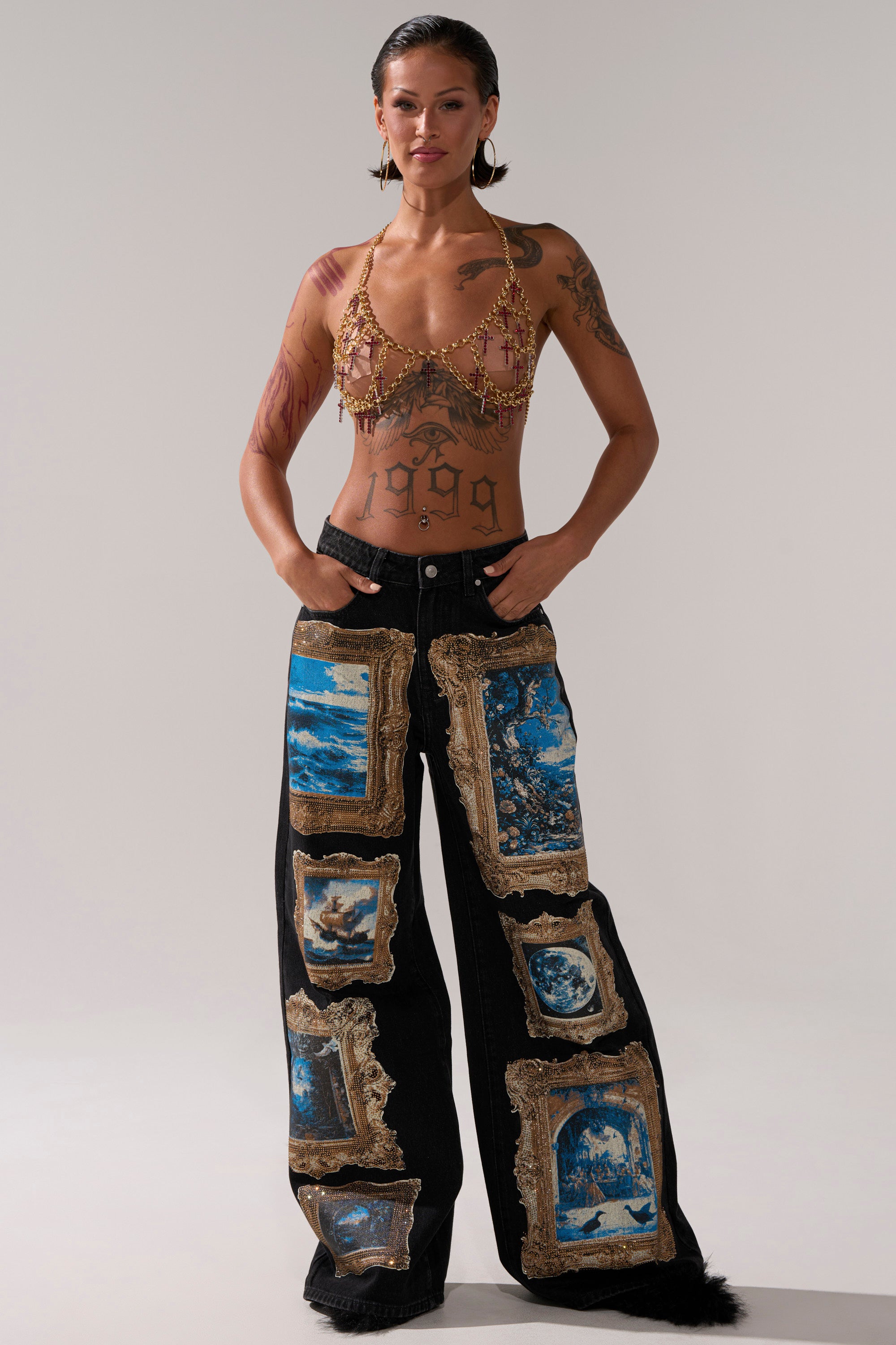 Wearing bold wide-leg black pants with blue and gold patches, a beaded bra top, and the HOLY CROSS BODY CHAIN, this confident individual showcases arm and torso tattoos against a plain light background that highlights their striking style.