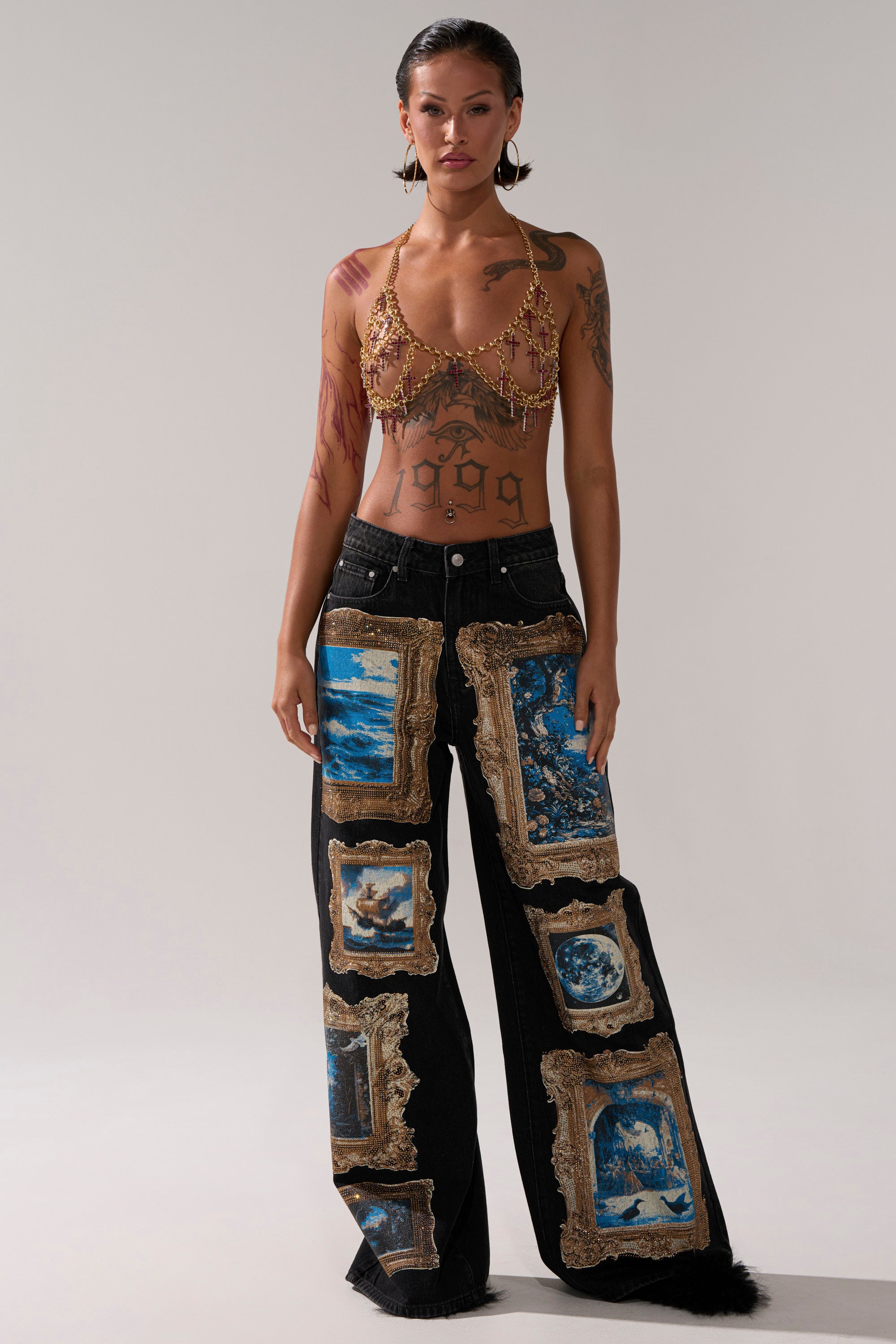 A person stands confidently in wide-leg black jeans with ornate artwork prints, a gold chain halter top, and the HOLY CROSS BODY CHAIN accentuating their body jewelry and tattoos on their torso and arms.