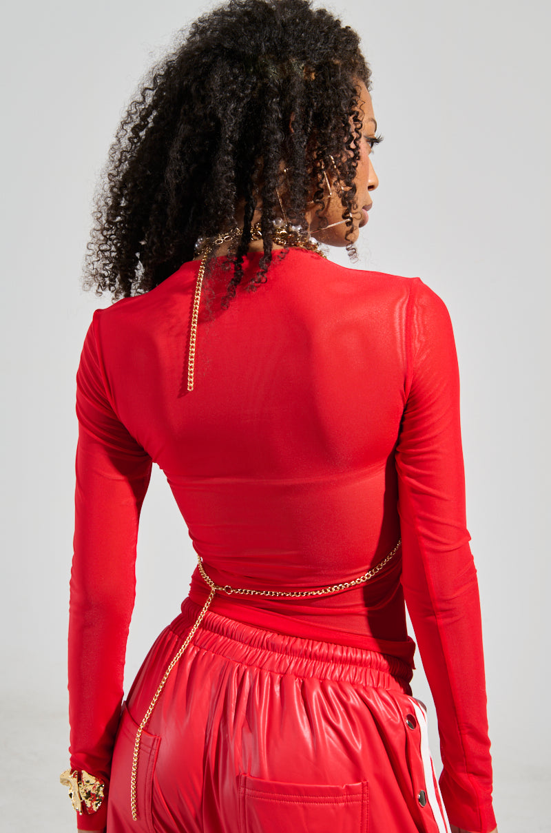 A person in a vibrant red outfit, consisting of a long-sleeved top and matching pants, stands with their back to the camera, highlighting their curly hair cascading down. They are elegantly adorned with the CLASSIC BODY CHAIN and a bracelet against the plain, light background.