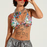 A person wearing large red sunglasses, a beaded top adorned with colorful beads, and metallic pants. The ROLL THE DICE CHARM BODY CHAIN elegantly graces their neck while their upper body features various tattoos, including the year "1999" and an eye design. They pose with one hand adjusting their sunglasses.