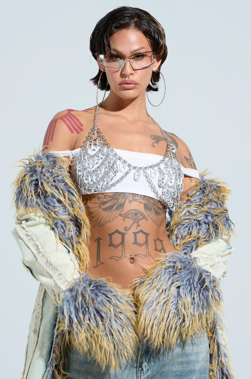 A person with short hair and sunglasses confidently poses wearing a fur-trimmed coat, silver-sequined adjustable halter neckline top, jeans, and the POPPIN TAGS BODY CHAIN. Tattoos cover their arms and torso, including a large "1999" on their stomach, against a plain background.