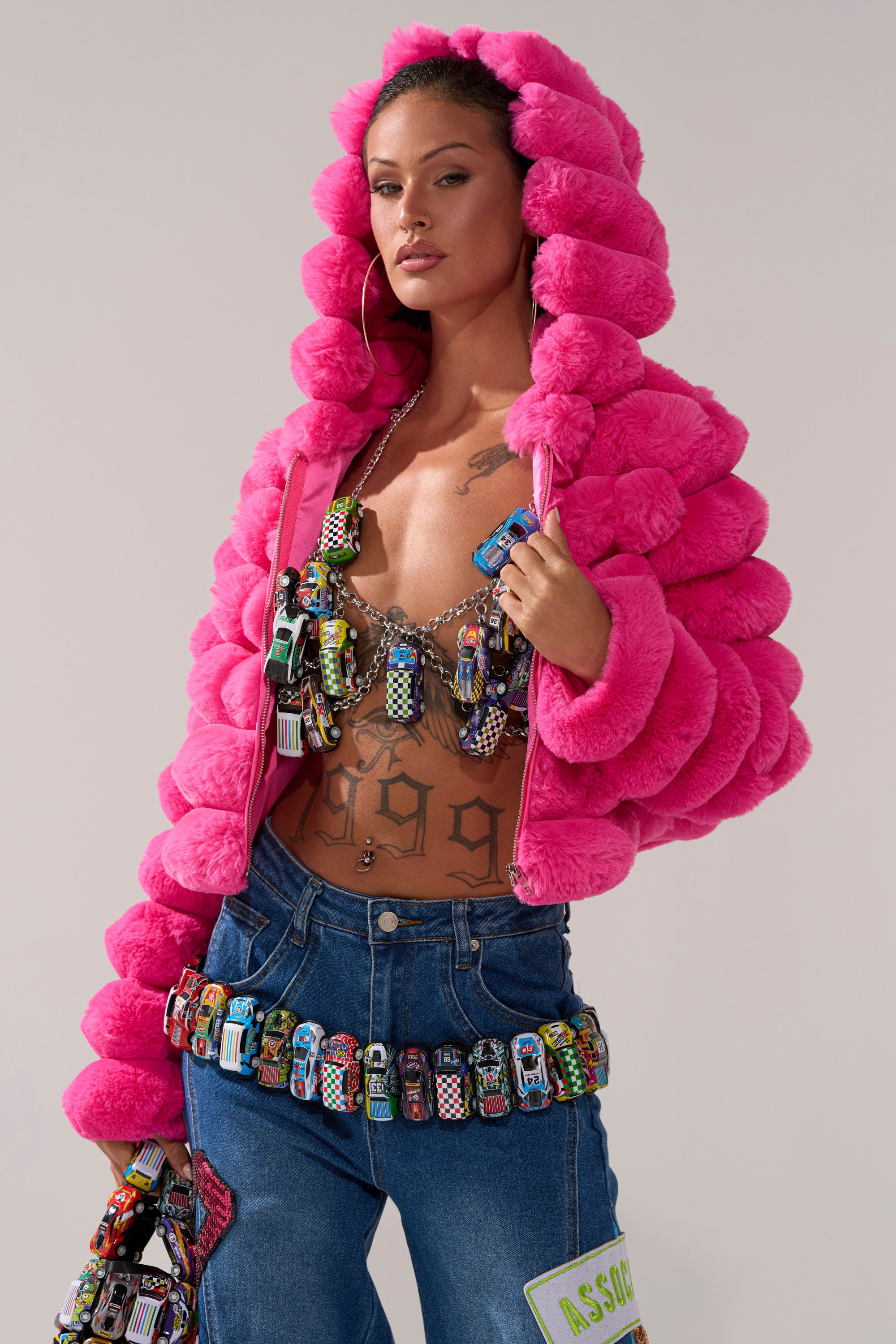 A woman wears the I GO FAST RACE CAR BODY CHAIN over a beaded bra, bright pink puffy fur jacket, blue jeans, and hoop earrings. She stands confidently with tattoos and colorful accessories against a plain background.