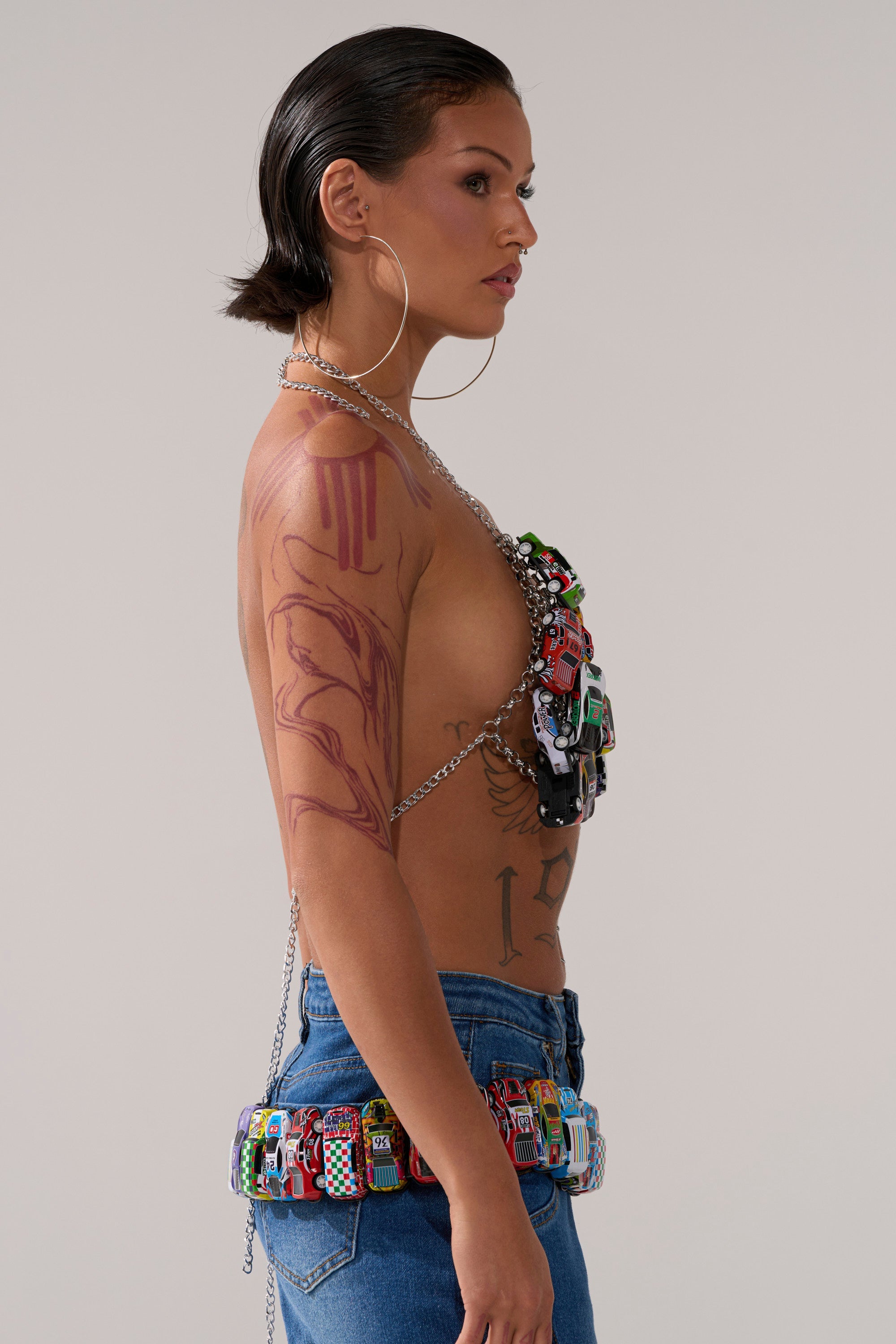 A woman with slicked-back dark hair, large hoops, and tattoos models the I GO FAST RACE CAR BODY CHAIN—a bikini-style accessory of colorful toy cars—paired with blue jeans. 3