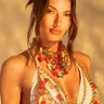 A woman with long brown hair in an updo wears a white bikini top adorned with colorful beads and chains, including the TASTE ME BODY CHAIN. Her statement necklace features vibrant, mixed stones. A softly lit background creates a warm atmosphere.