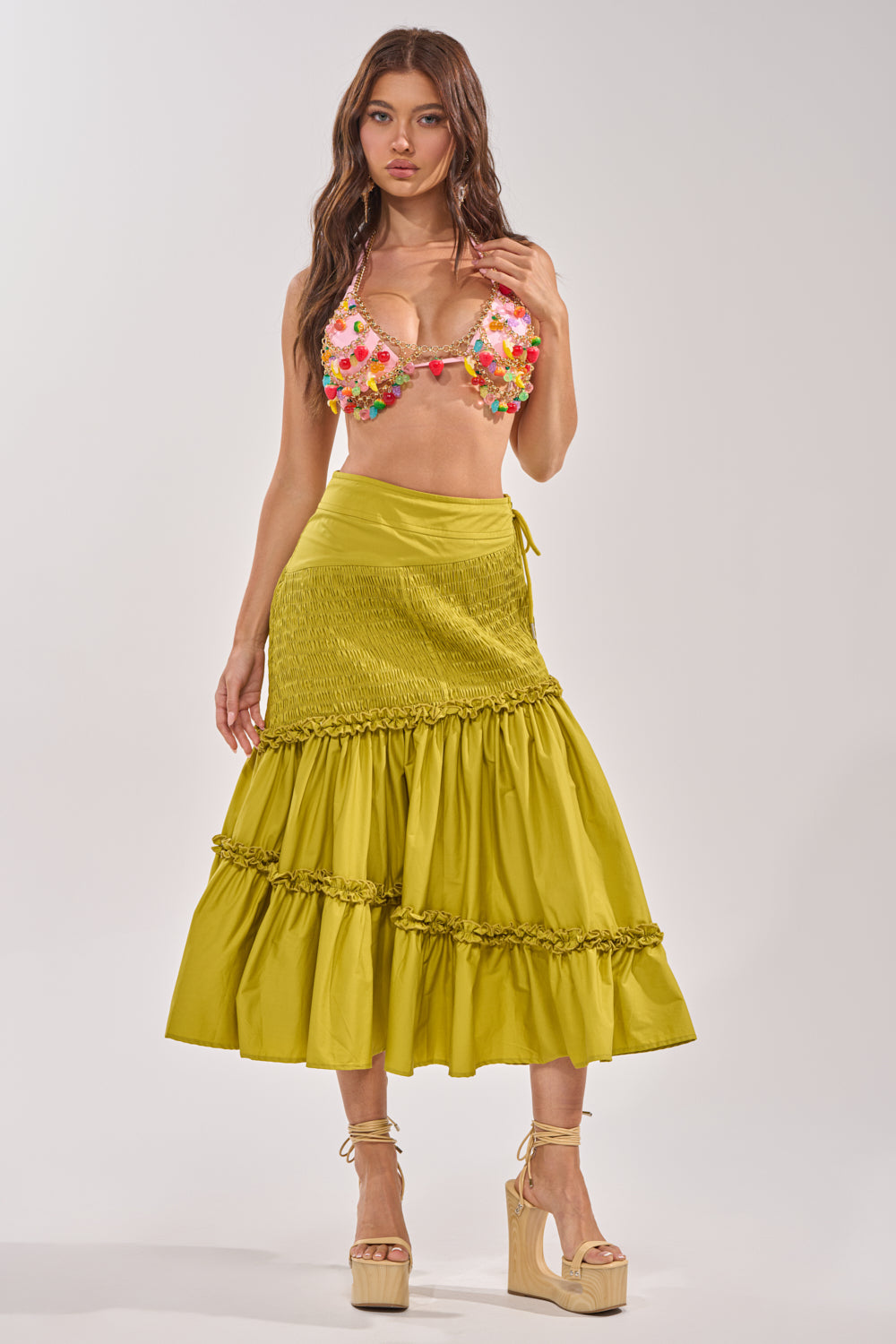 A woman stands against a plain background wearing a colorful floral bikini top, a high-waisted yellow tiered skirt, beige platform sandals with ankle ties, and the TASTE ME FRUITY BODY CHAIN for a playful festival touch. 5