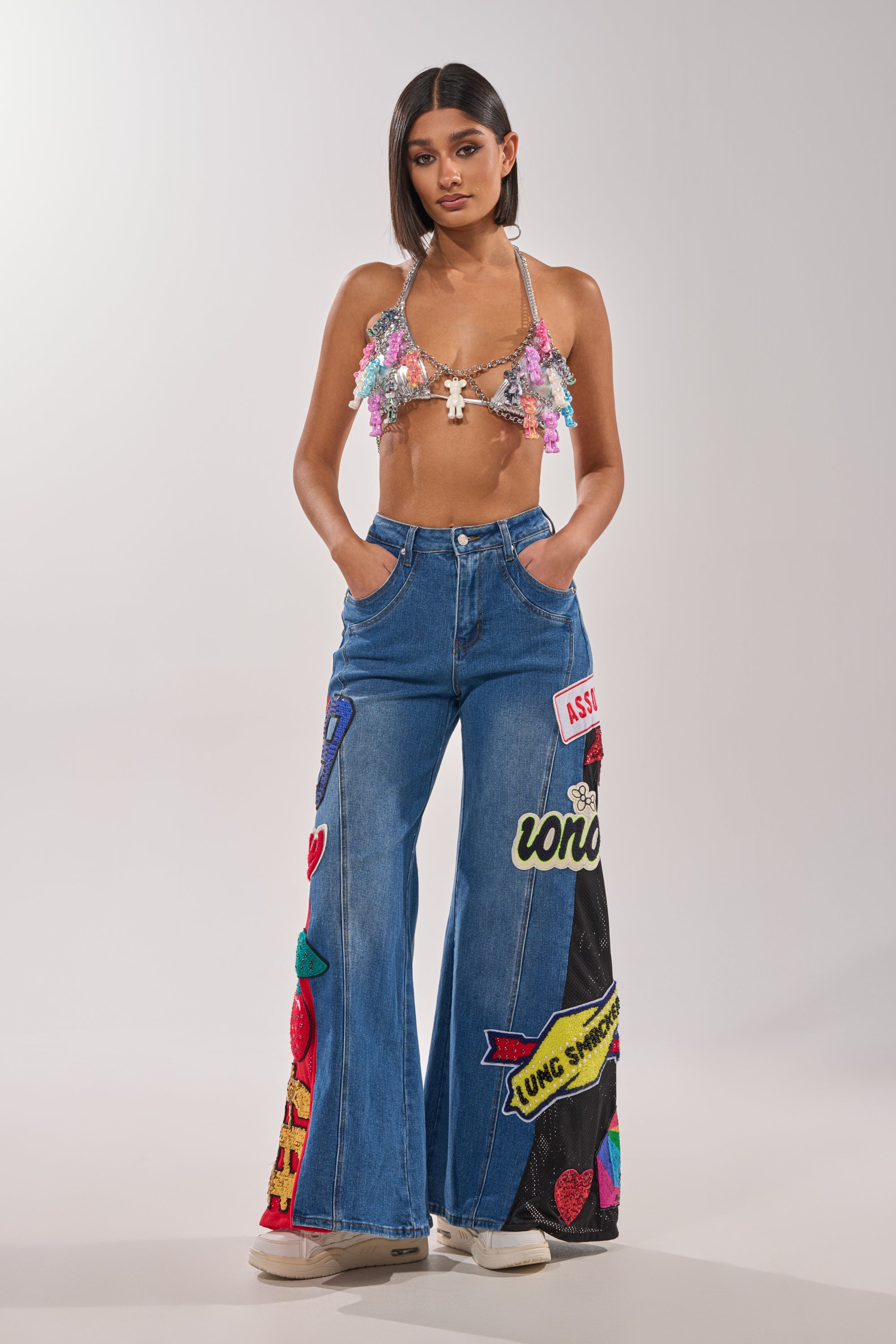 A woman stands facing forward in a Y2K-inspired look, wearing a sparkling sequin bralette, blue wide-leg jeans with colorful patches and text, white sneakers, and the BEARY CUTE BODY CHAIN. Her hands rest in her pockets. 5