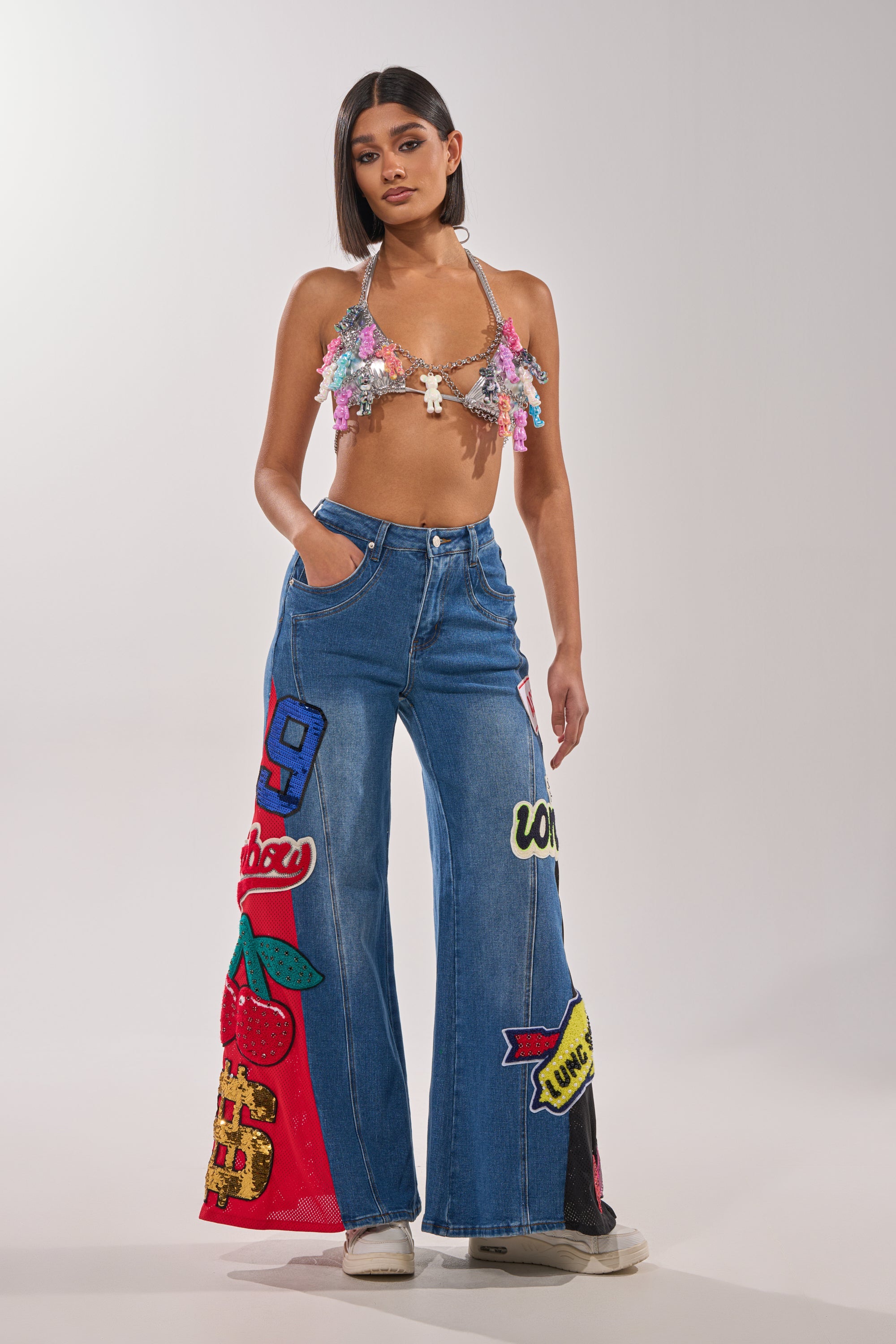 A woman stands against a plain light background in flared blue jeans with colorful patches, a floral sequin bikini top, white sneakers, and the BEARY CUTE BODY CHAIN, channeling a Y2K-inspired look. 6