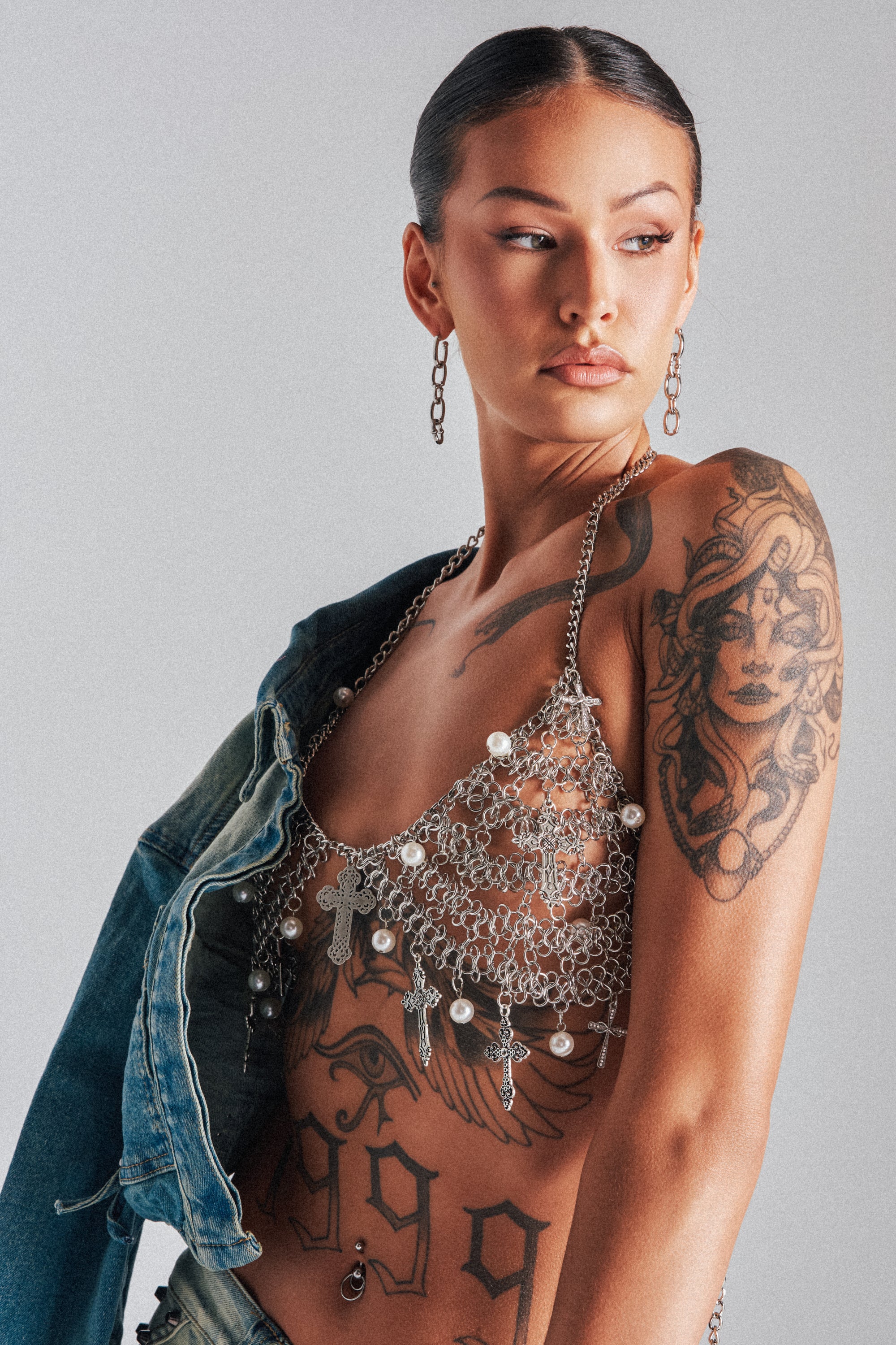 A woman with slicked-back hair models the CHARMED LIFE BODY CHAIN, a layered silver-tone chain and pearl top, with one arm in a denim jacket. Tattoos adorn her arm and side as she poses against a plain light background.