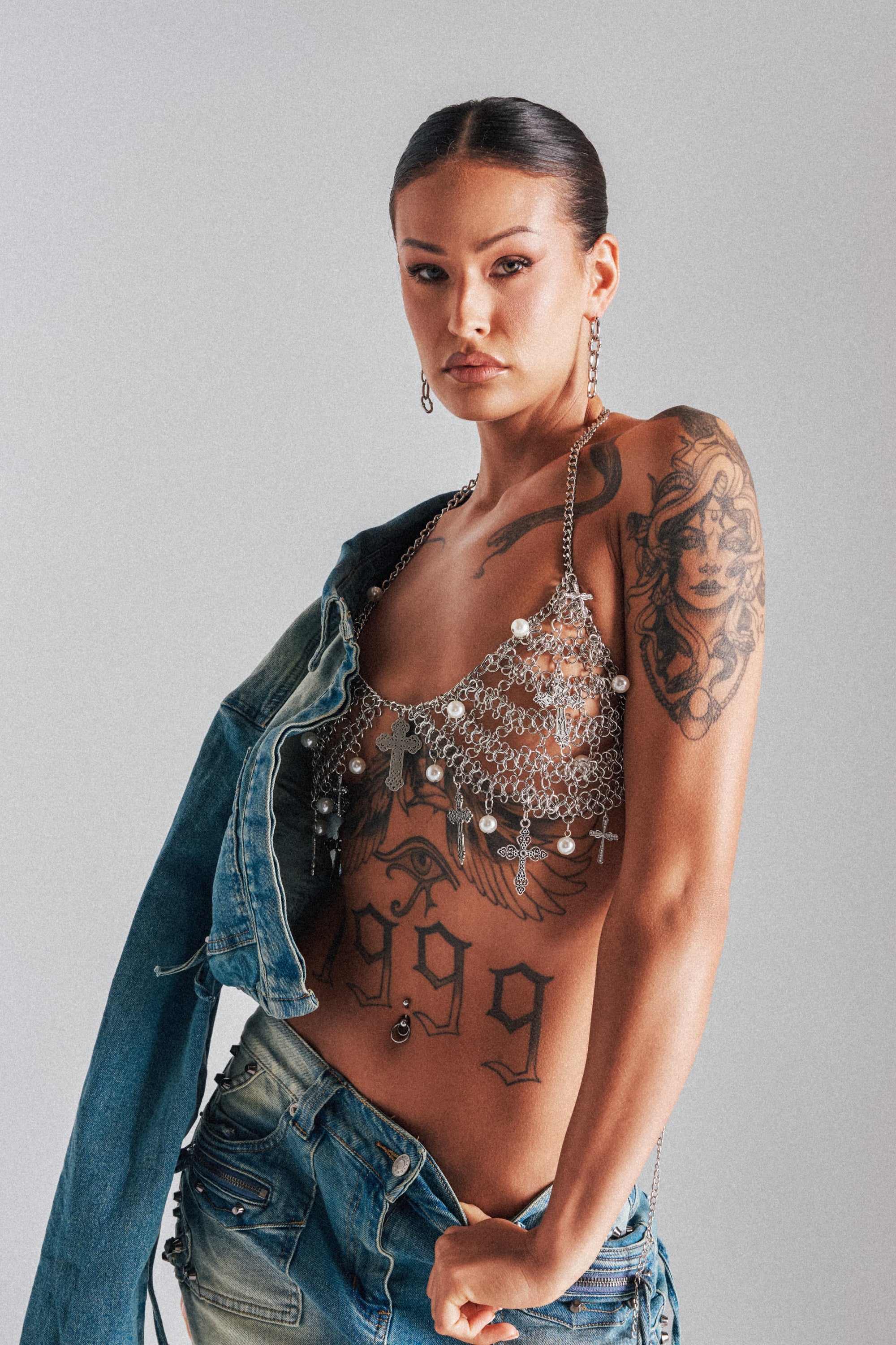 A woman with tattoos poses confidently wearing the CHARMED LIFE BODY CHAIN layered over a pearl top, denim jacket off one shoulder, and low-rise jeans. She has slicked-back hair, dangling earrings, and a bold "1999" torso tattoo. 1