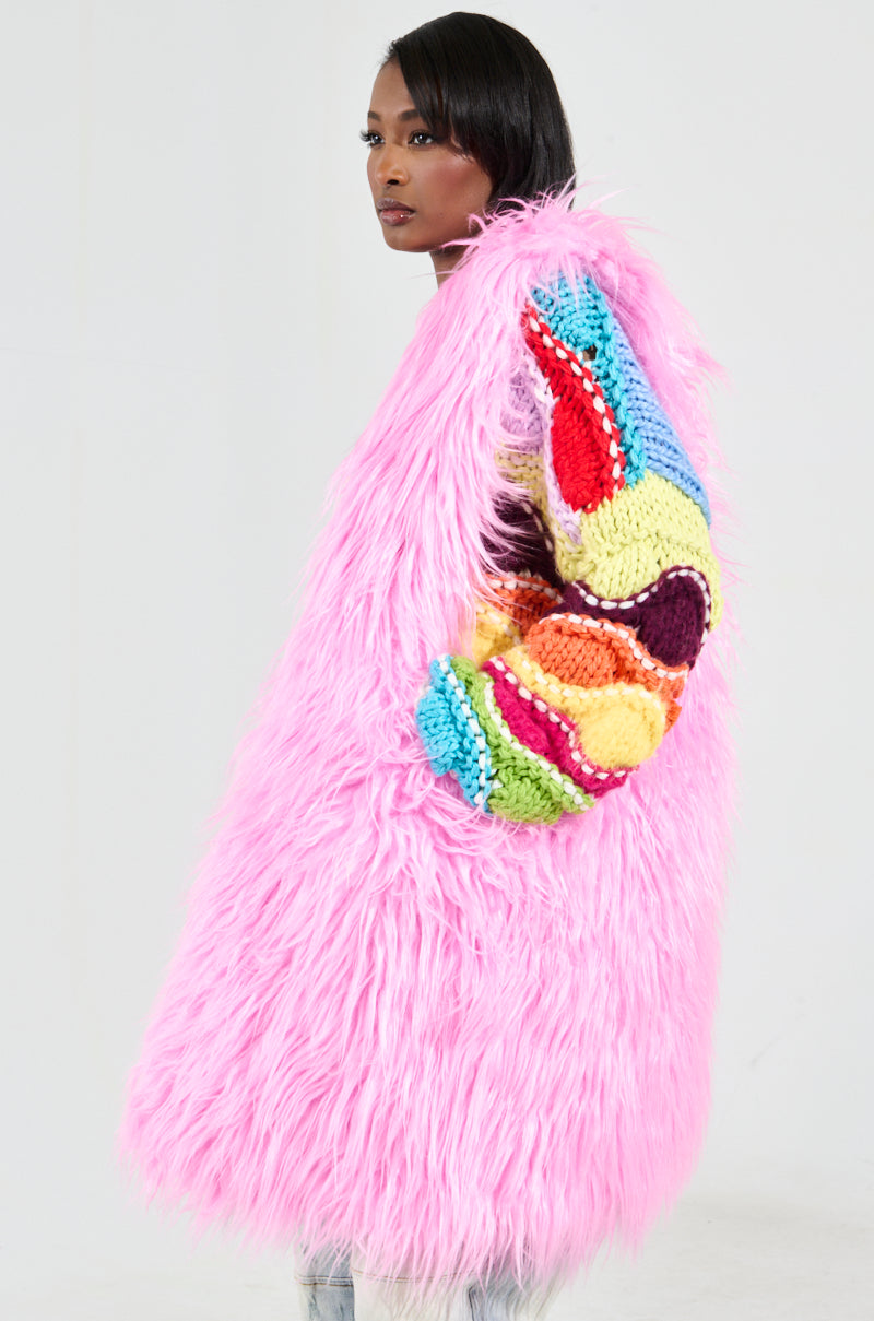 The person is dressed in a long, fluffy pink coat adorned with colorful knitted details on the shoulder and sleeve. They are carrying the "I LOVE YOU Faux Fur Oversized Tote" in bubblegum pink, adding a playful touch. The background is plain and light-colored.