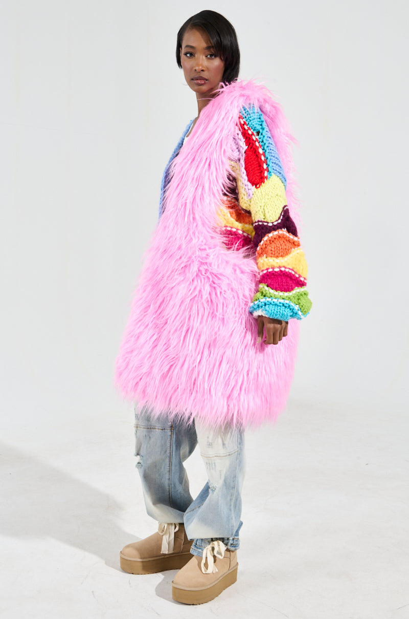 A person dressed in a striking pink faux fur coat featuring multicolored, textured sleeves is posed against a simple background. They carry the I LOVE YOU FAUX FUR OVERSIZED TOTE in bubblegum pink, which brings an extra touch of style to their outfit. Their ensemble is balanced with loose-fitting jeans and beige platform shoes, while their short hair and neutral expression enhance the fashionable look.