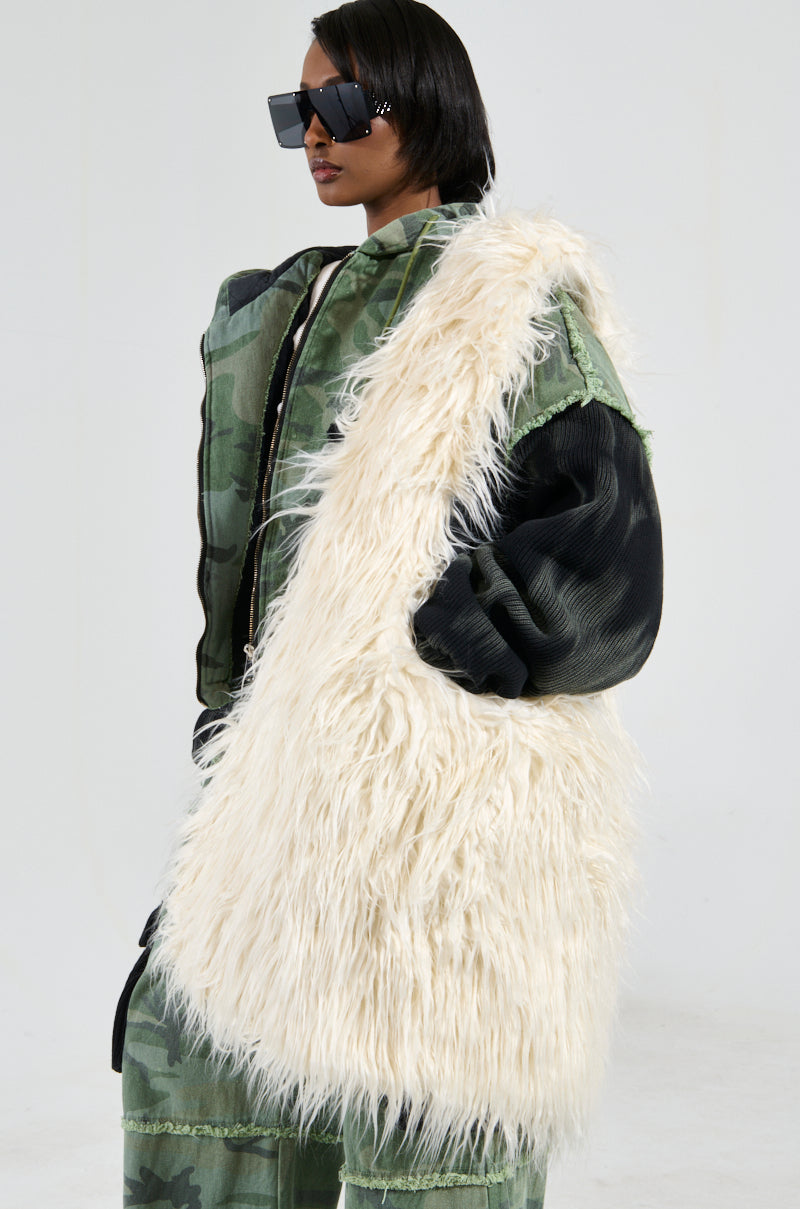 A person wearing oversized sunglasses, a green camouflage jacket, and loose-fitting pants stands confidently with the SHAGGADELIC FAUX FUR OVERSIZED TOTE. Draped in a large, white, fluffy coat featuring spacious interior pockets, they pose against a plain light-colored background.