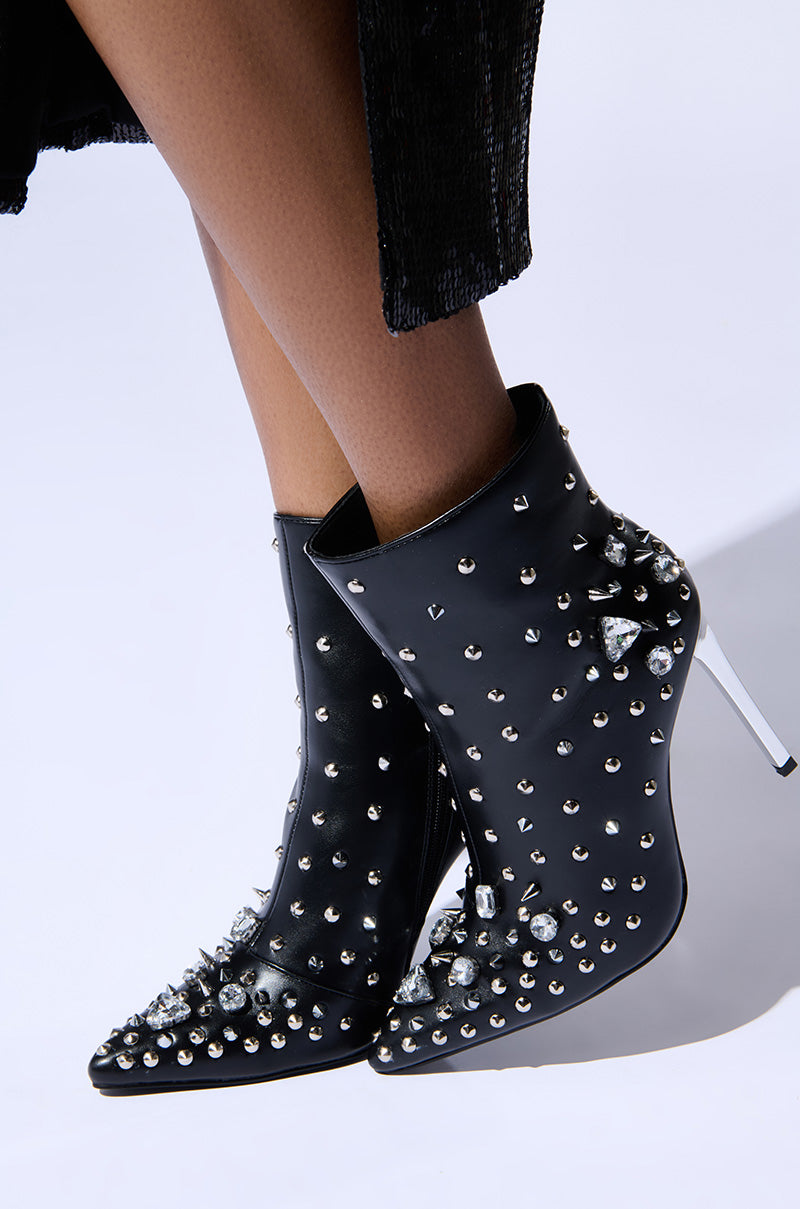 Introducing the AZALEA WANG OAKLEE BLACK STILETTO BOOTIE, a stunning pair of ankle boots featuring silver spikes and studs. Crafted in glossy faux leather, these striking boots are paired with a black textured skirt, all set against a plain white background to highlight their unique design.