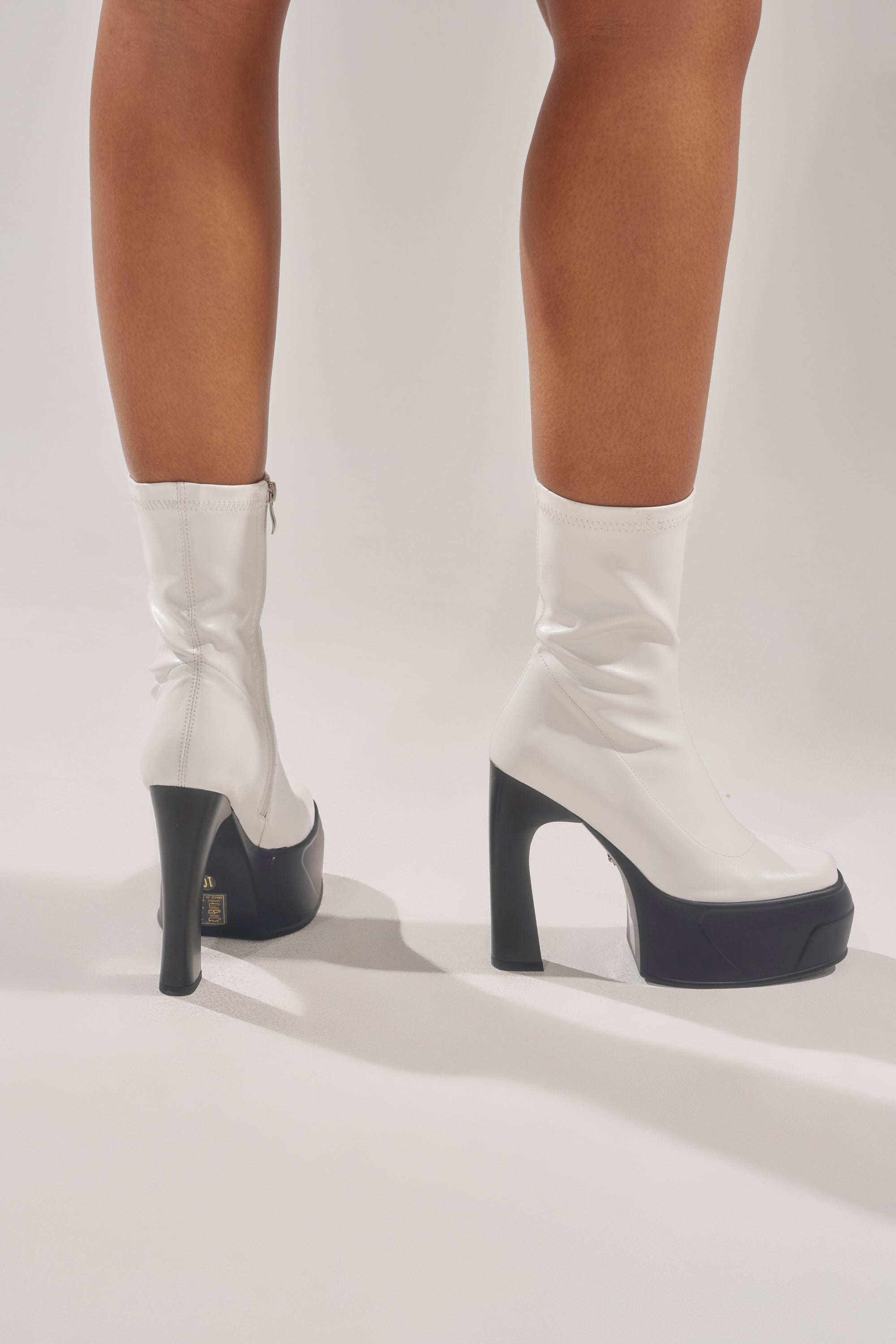 A person wears the AKIRA GOLD OBSIDIAN WHITE CHUNKY PLATFORM BOOTIE with shiny white faux leather, chunky platform heels, and an ankle zip, standing on a light surface. Only their lower legs and feet are visible. 0