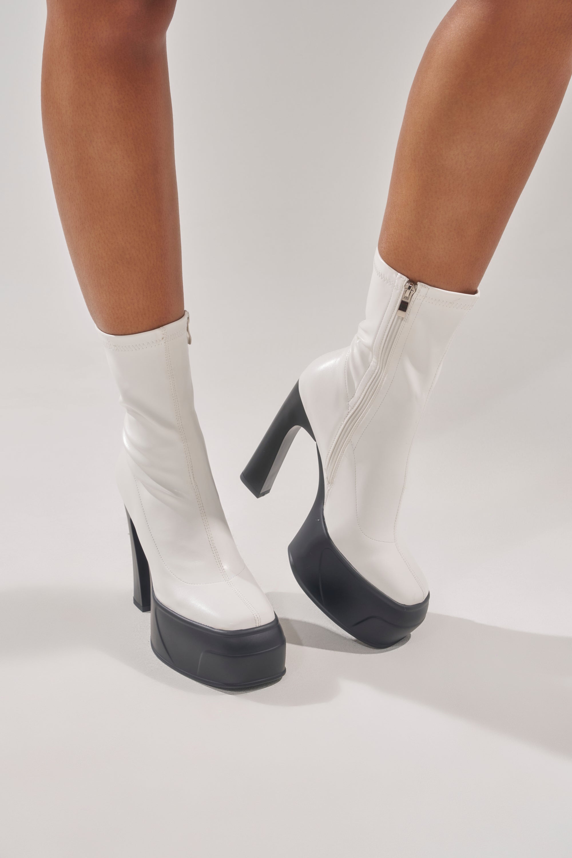 A person stands on a white background wearing the AKIRA GOLD OBSIDIAN WHITE CHUNKY PLATFORM BOOTIE, featuring white faux leather, high black chunky heels, and side zippers for a bold, modern look. 1