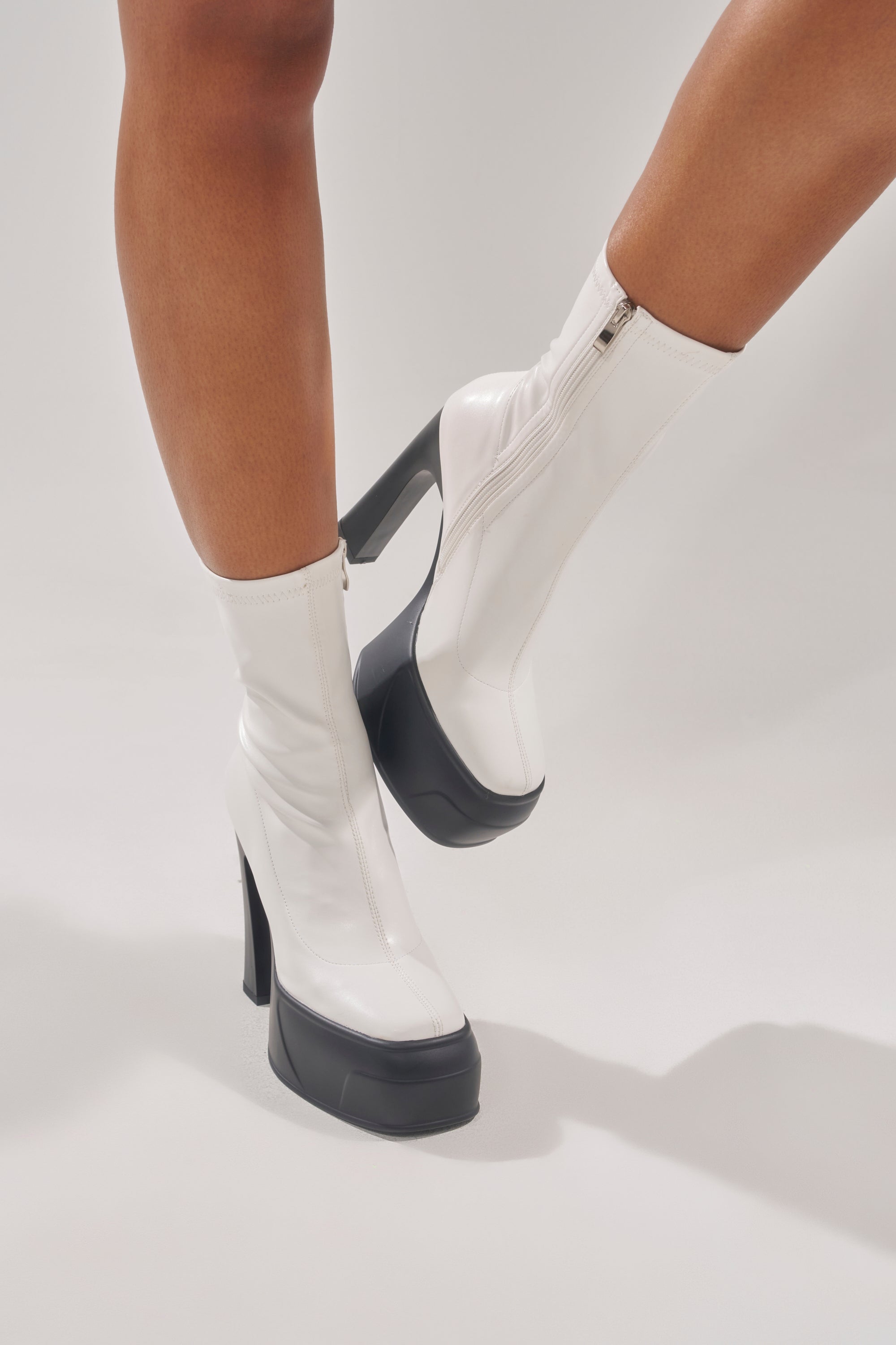 Someone wearing the AKIRA GOLD OBSIDIAN WHITE CHUNKY PLATFORM BOOTIE, featuring white faux leather, black platform soles, and chunky heels, stands against a plain light background. 2
