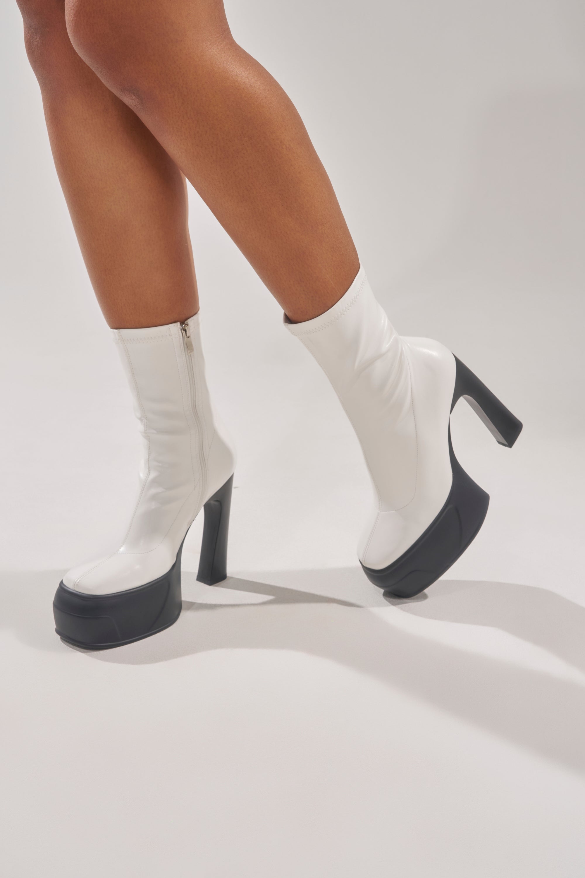 Wearing the AKIRA GOLD OBSIDIAN WHITE CHUNKY PLATFORM BOOTIE, a person stands on a light gray surface. Only the lower legs and feet are visible, showcasing the bold style of these white chunky platform booties. 3