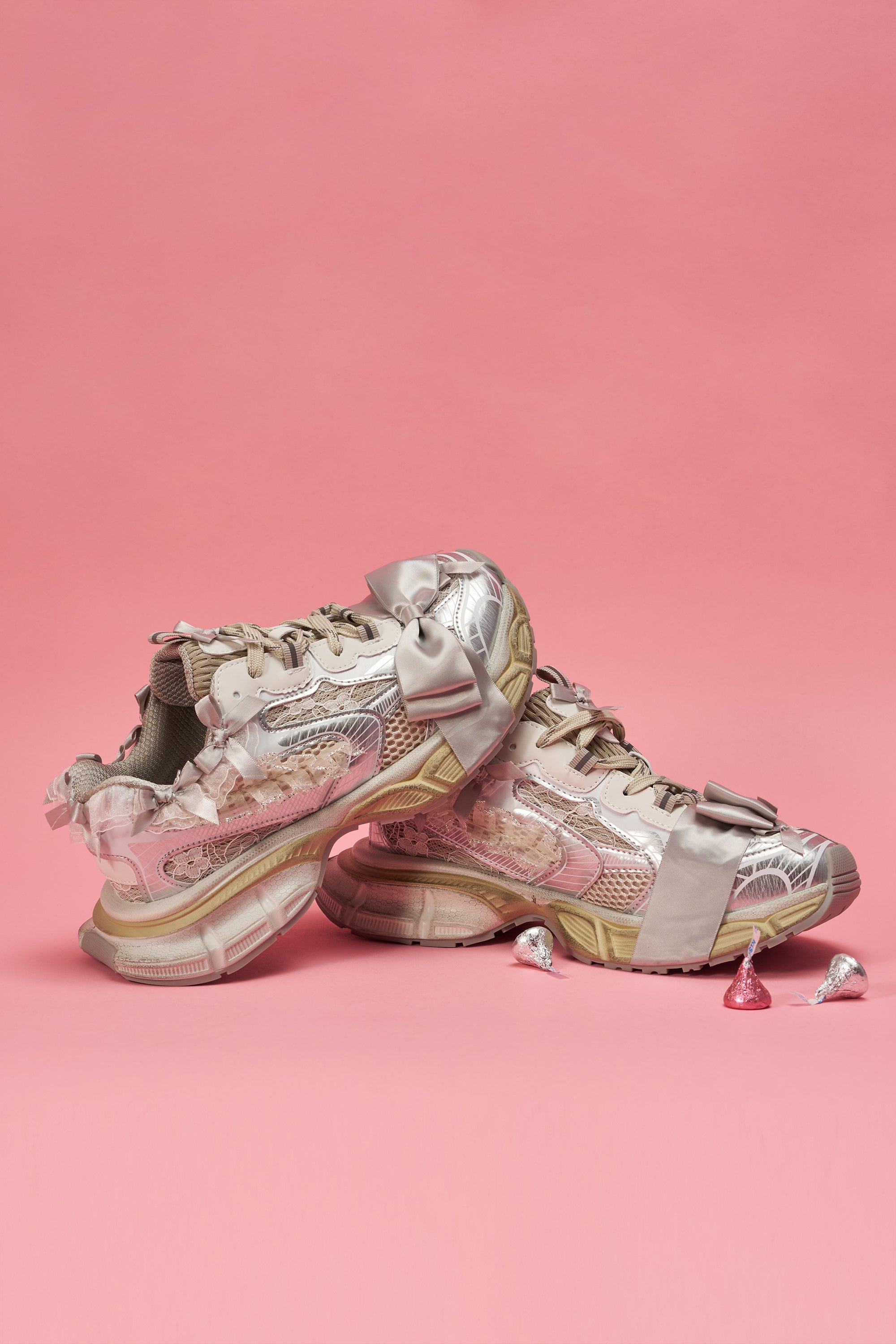 Two AZALEA WANG OCTAVIA White Bow Embellished Chunky Sneakers with shiny details and large satin bows are stacked on a pink background, with three wrapped Hershey’s Kisses chocolates in front, emphasizing their cute, girly style. 0
