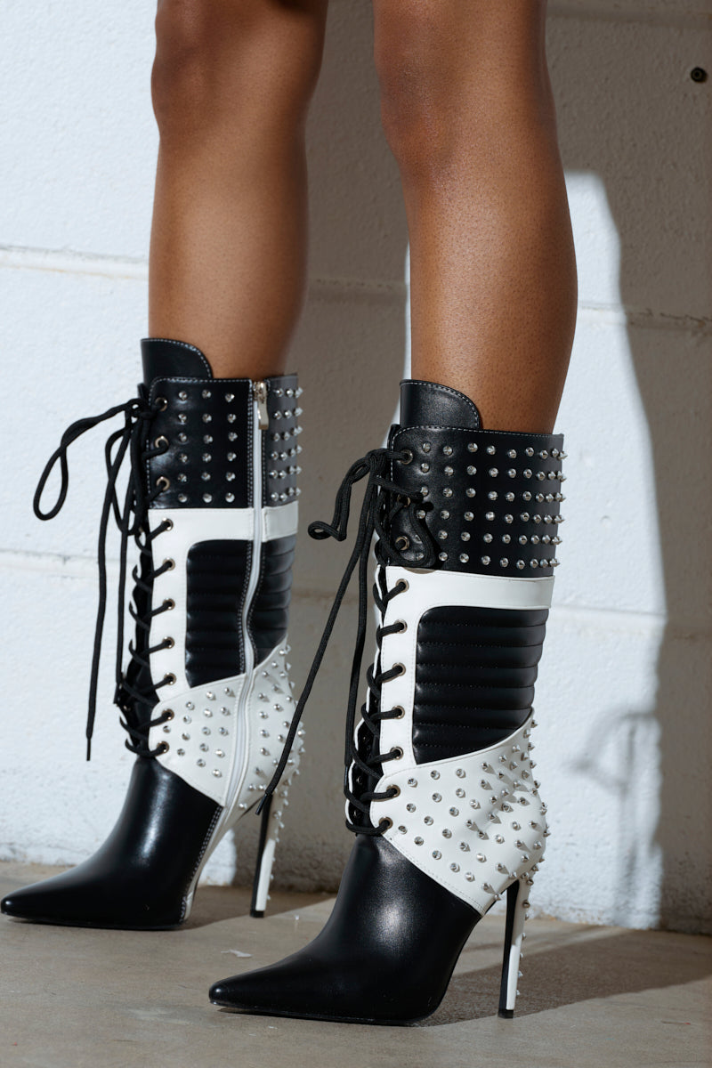 Person wearing the AZALEA WANG OCTOBI BLACK AND WHITE STILETTO BOOT has stylish knee-high boots with a black-and-white design, pointed toe, and faux leather upper. Stiletto heels with studded details add flair to the lace-up fronts against a white background.