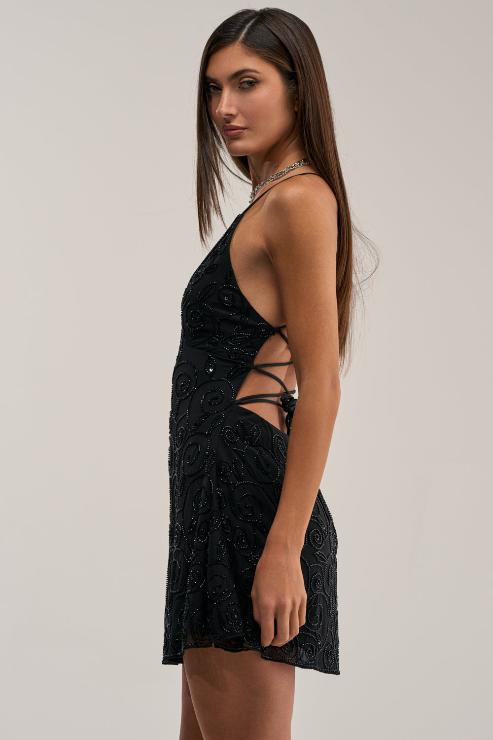 A woman with long straight brown hair stands in profile against a plain light background, wearing the TEMPTED TONIGHT EMBELLISHED MINI DRESS—a sleeveless black mini dress with beading, side lace-up details, and a patterned texture.