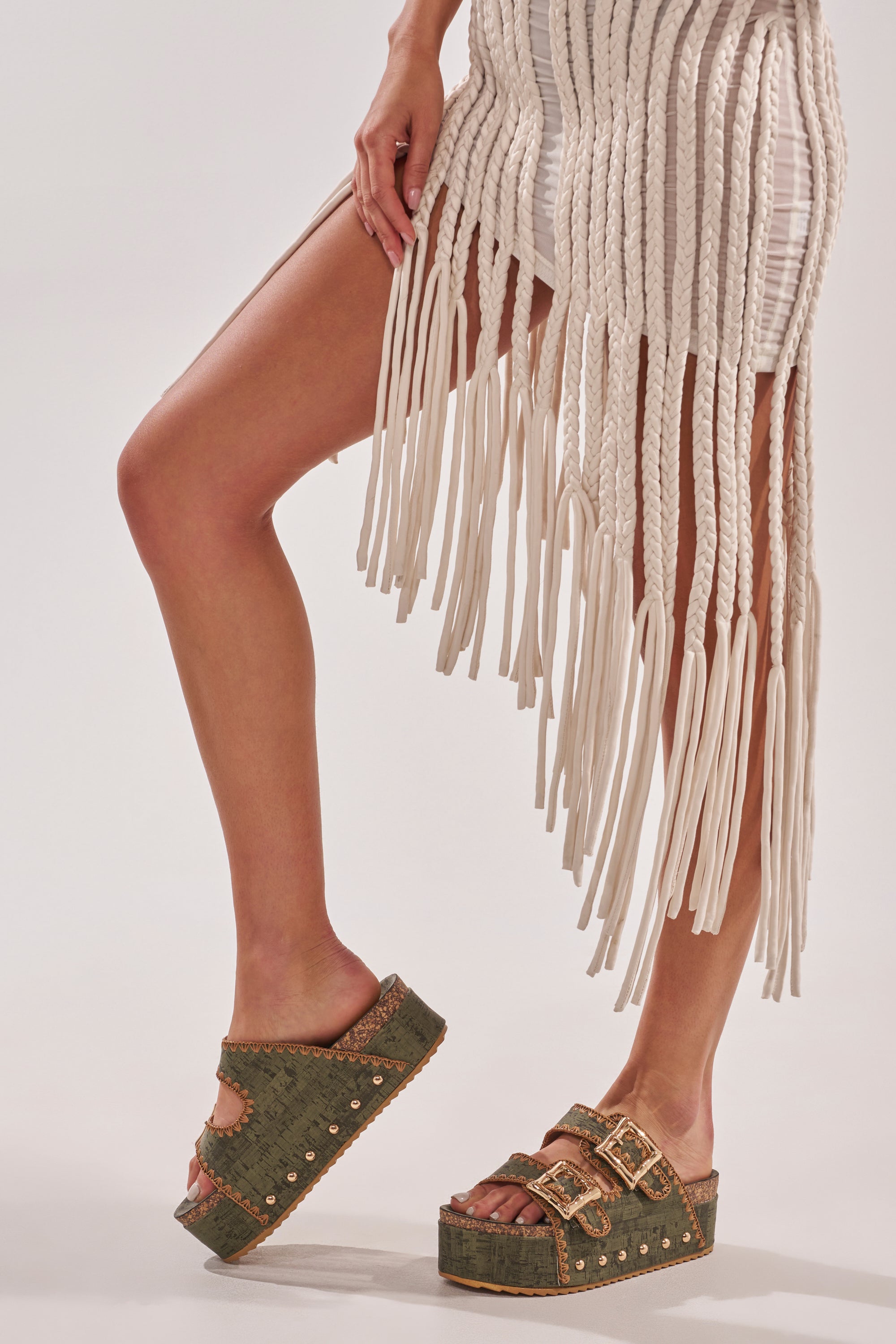 A person stands against a plain background, showing only their legs and a white dress with long fringe while wearing AZALEA WANG OHKAY GREEN FLATFORM STUD EMBELLISHED SLIP ON SANDALS. 0