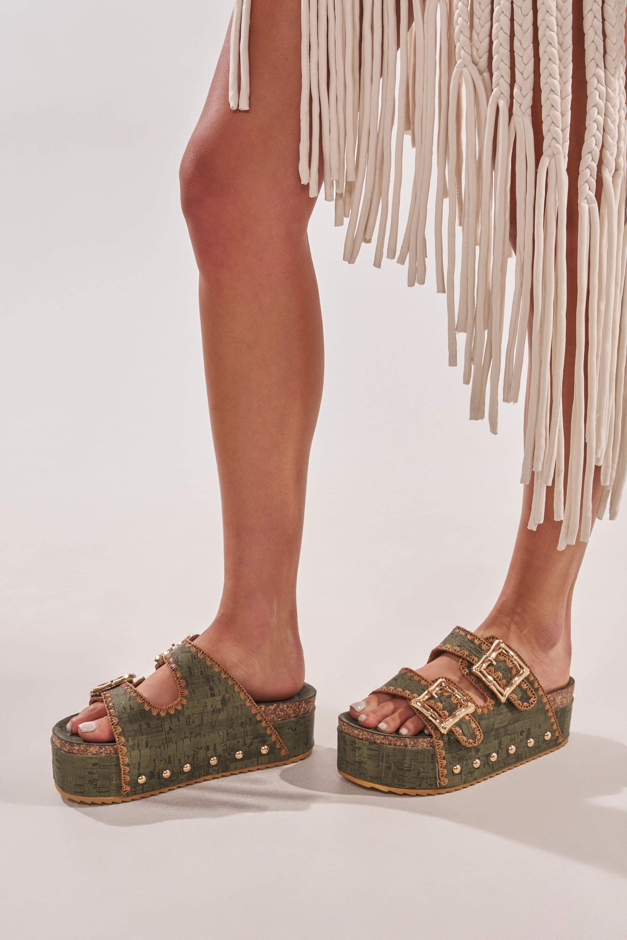 A person wearing a white fringed skirt and AZALEA WANG OHKAY GREEN FLATFORM STUD EMBELLISHED SLIP ON SANDALS stands on a light-colored surface. 2