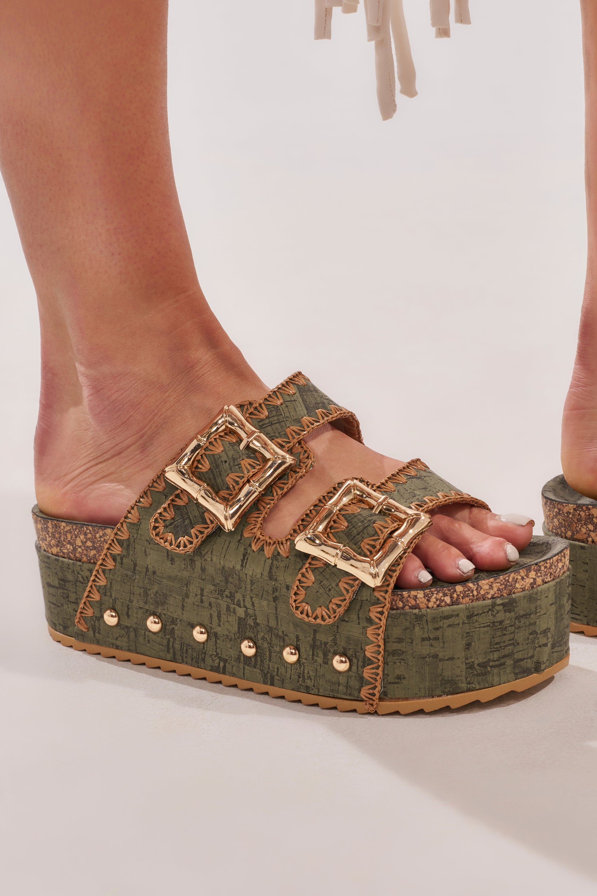 Close-up of the AZALEA WANG OHKAY Green Flatform Stud Embellished Slip On Sandal, featuring olive green faux leather, two large gold buckles, gold stud accents, a cork flatform sole, textured finish, and visible stitching. 7