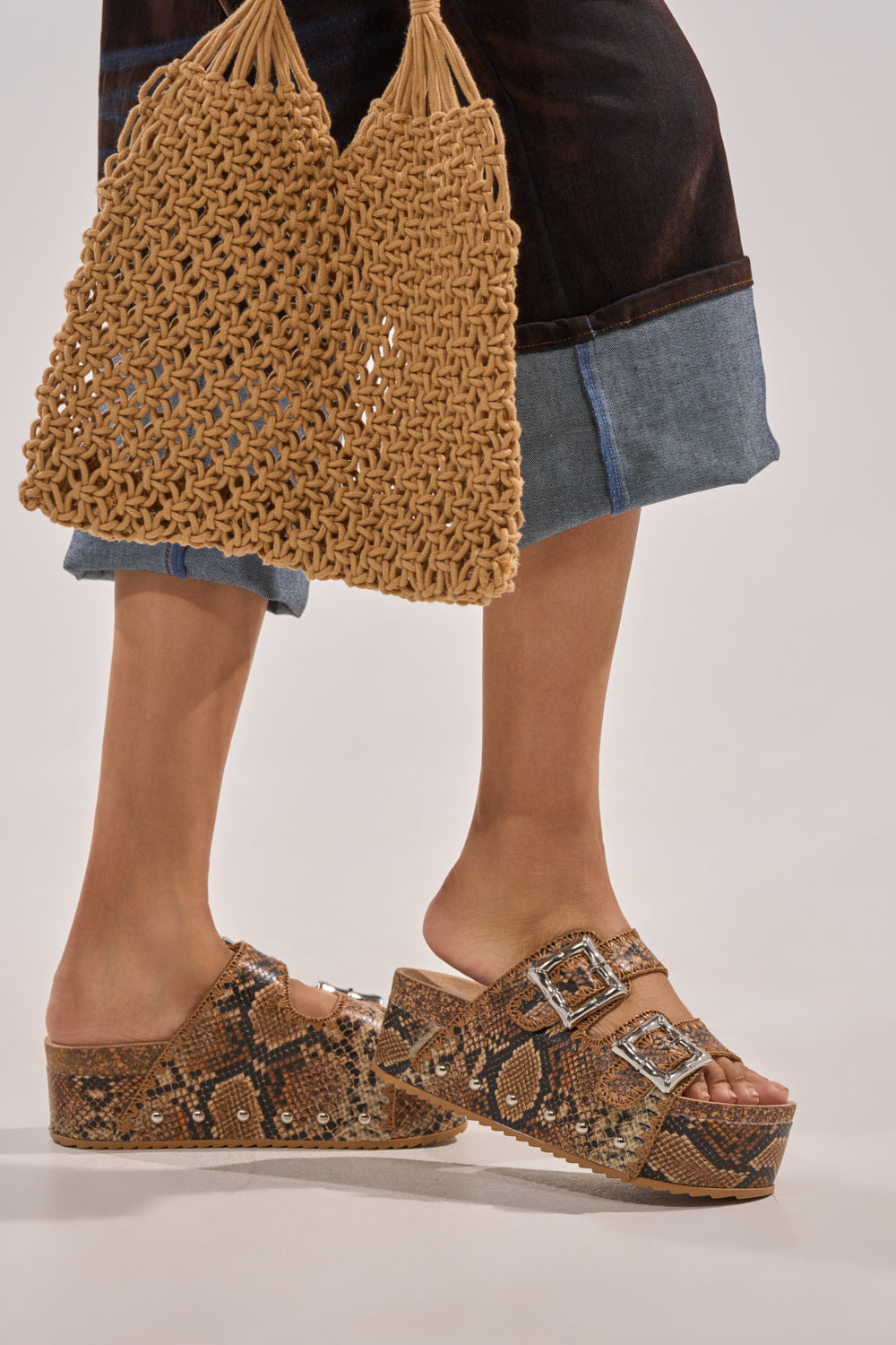 Wearing AZALEA WANG OHKAY SNAKE FLATFORM STUD EMBELLISHED SLIP ON SANDALS with chunky soles and buckles, paired with a woven tan bag and a dark skirt with a denim hem. 0