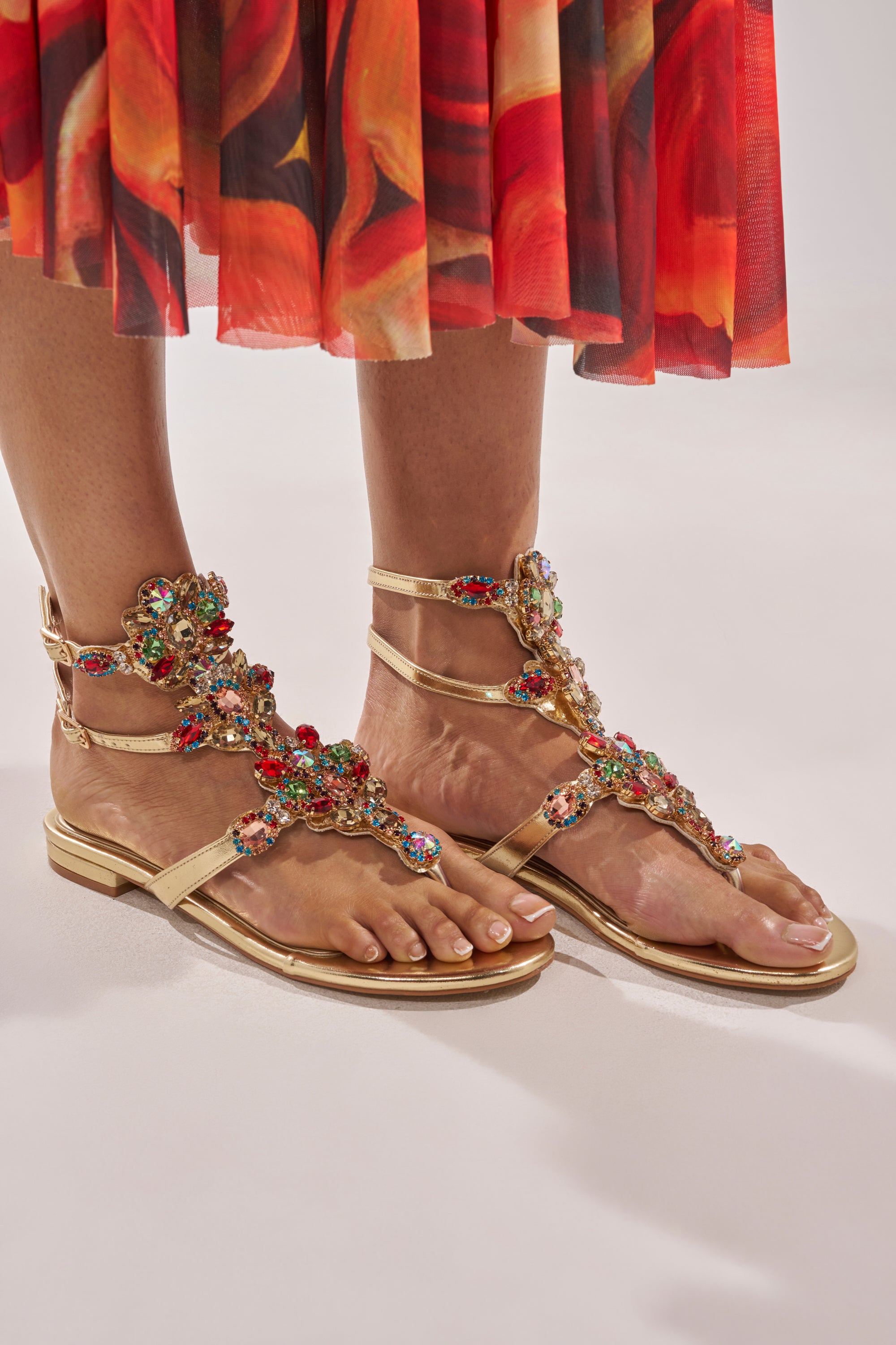 A person wearing AZALEA WANG OLISA GOLD RHINESTONE EMBELLISHED FLAT SANDALS paired with a vibrant red and orange patterned skirt, with the photo highlighting their feet and lower legs. 0
