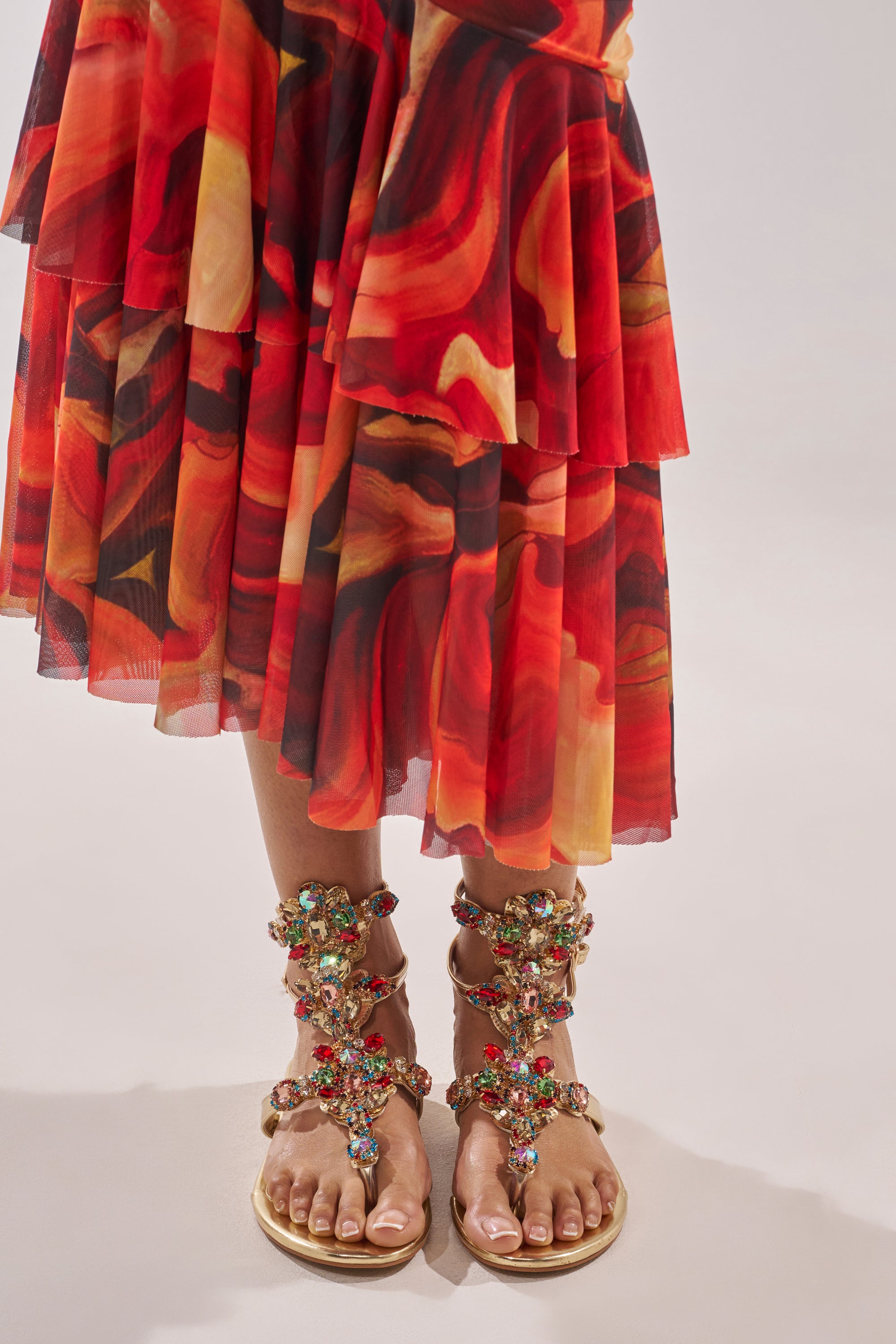 A person stands on a light surface, wearing a vibrant red and orange patterned skirt and the AZALEA WANG OLISA GOLD RHINESTONE EMBELLISHED FLAT SANDAL adorned with sparkling gemstones. 1